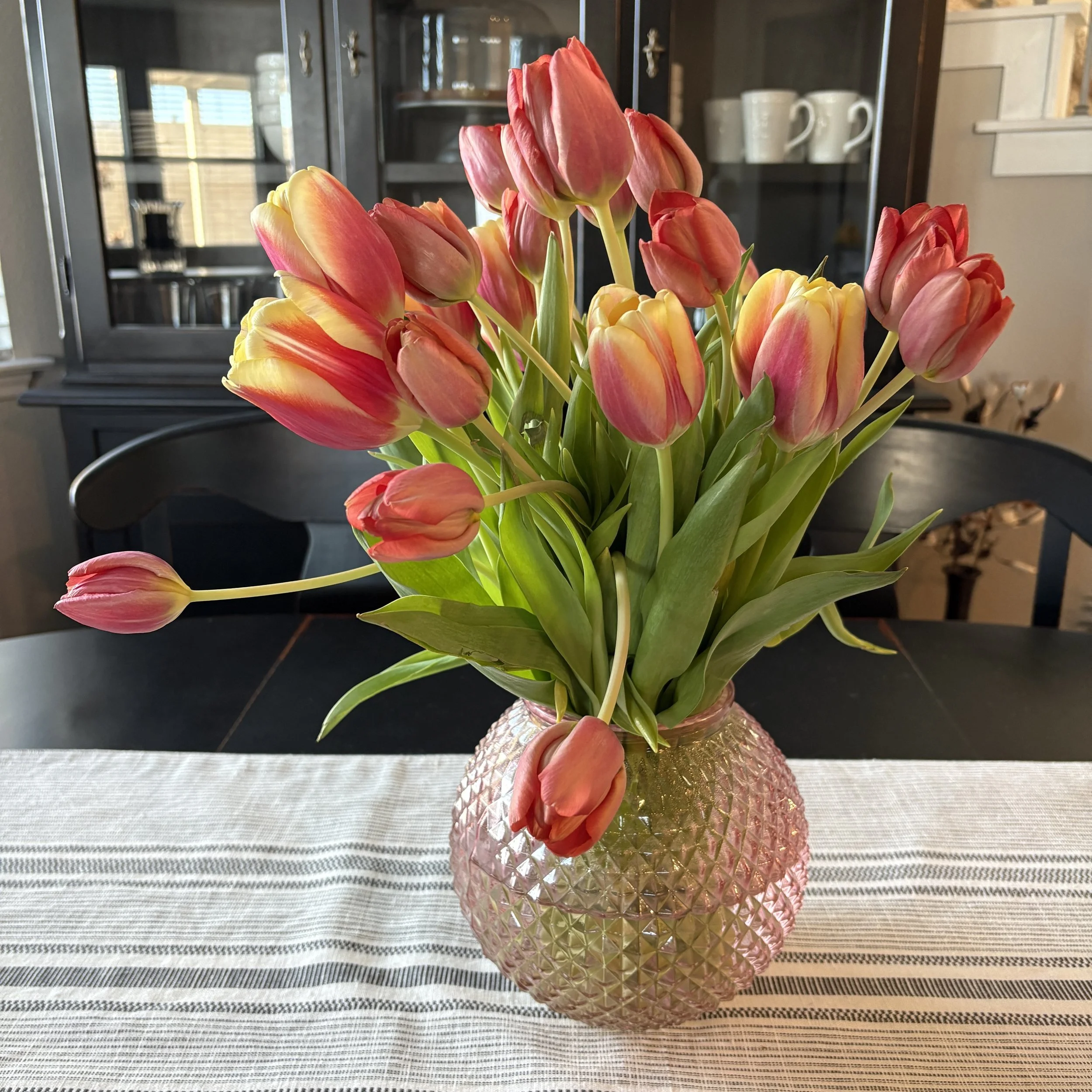Pink and yellow tulips arranged in a textured pink vase on a striped table runner in a dining area.
