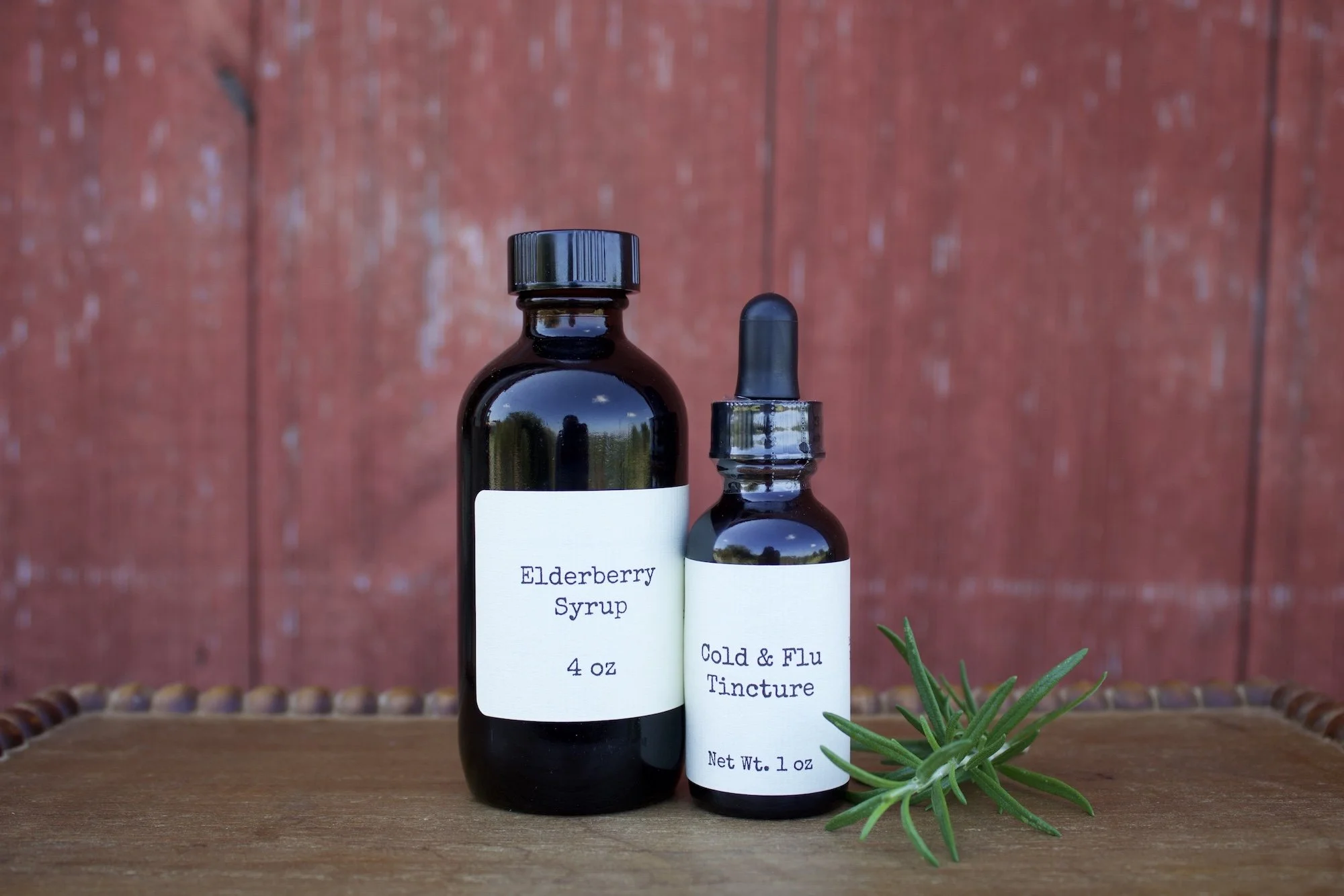 Two dark glass bottles in front of a red wooden wall with a sprig of rosemary beside them