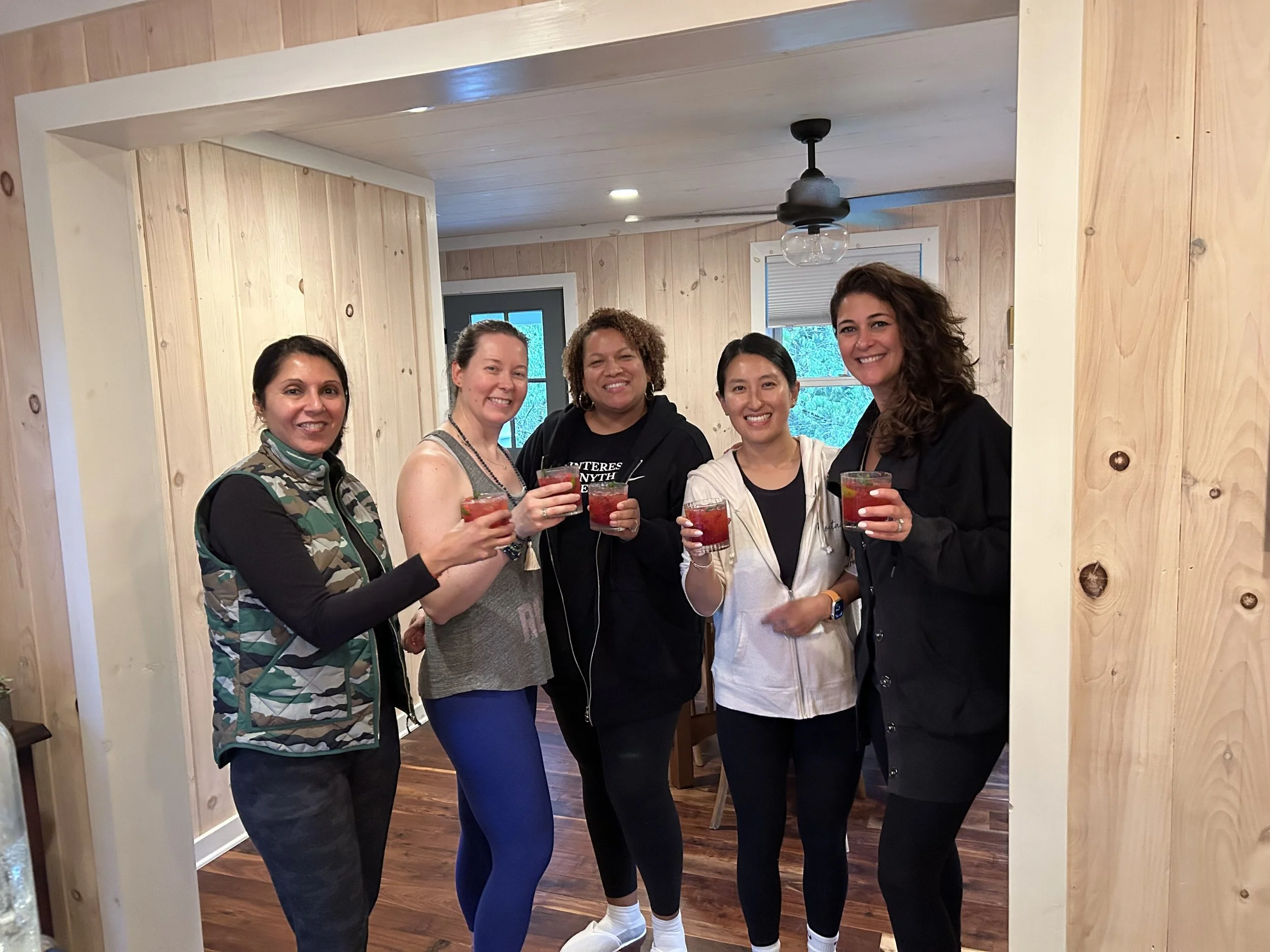 Group of smiling women holding red drinks standing in a natural wood room