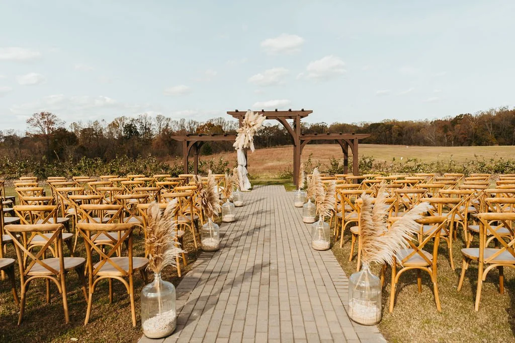 Brick path lined with wooden chairs facing wooden archway against a backdrop of a field bordered with trees