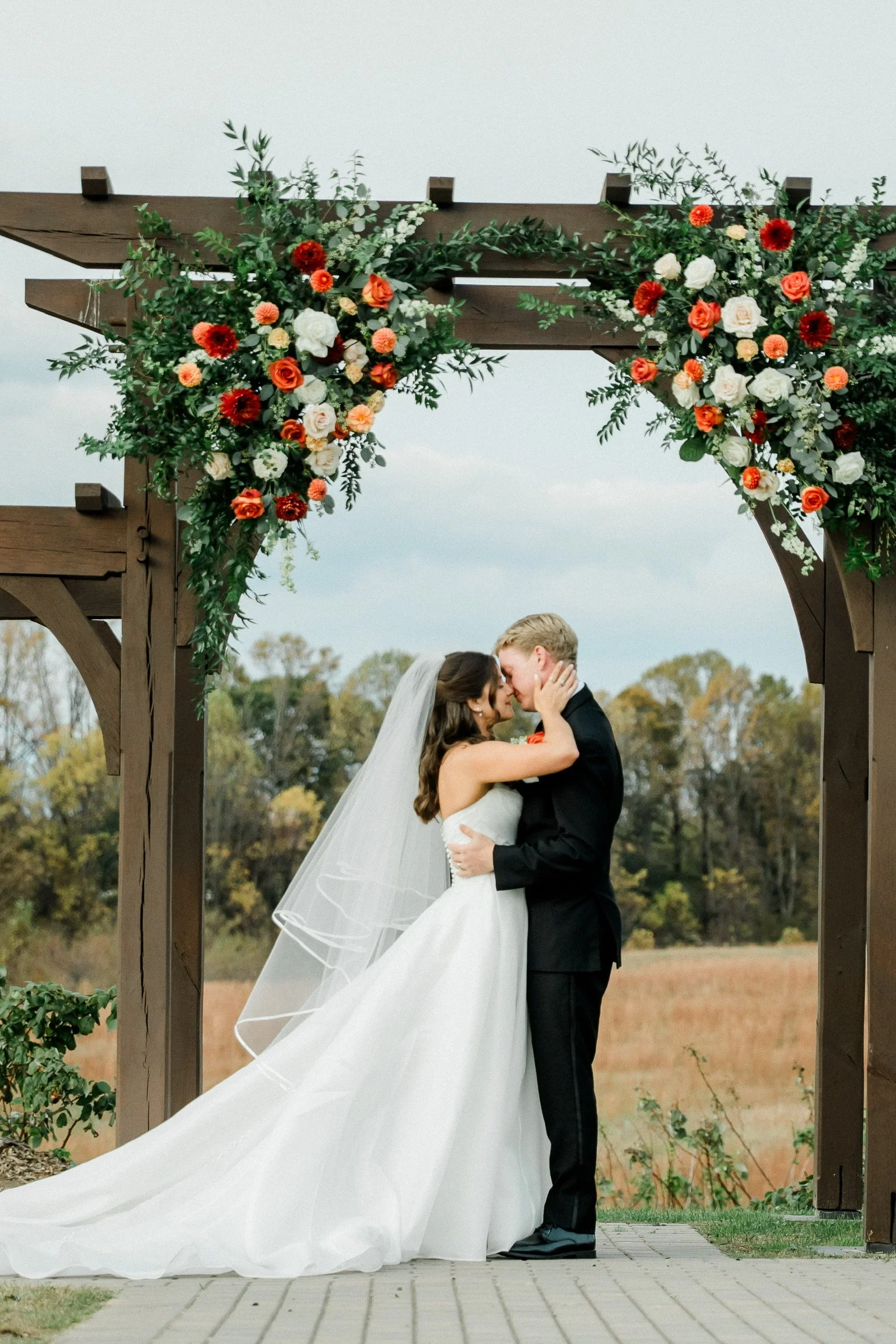 Woman in a white dress and veil and man in a black suit kiss under an archway decorated with flowers