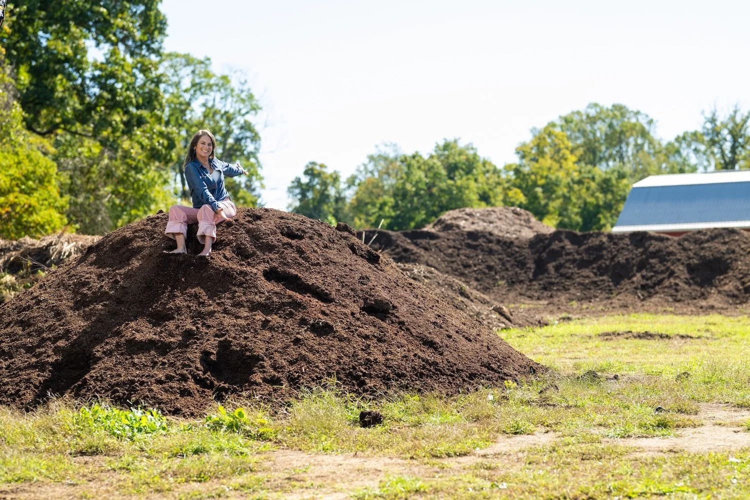 Woman in a blue shirt and pink pants sitting on top of a pile of dirt with arms outspread