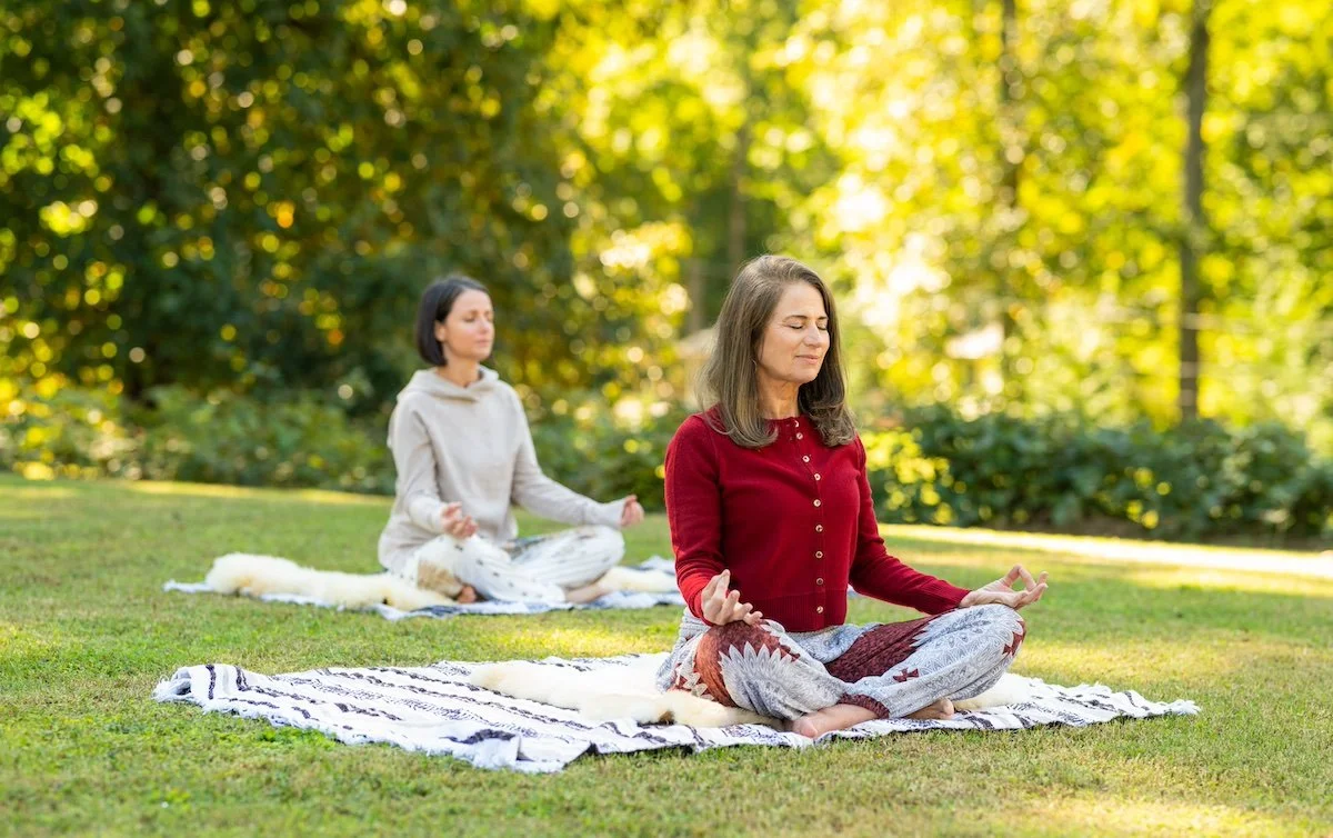 Two women sitting on blankets outside meditating with greenery in background