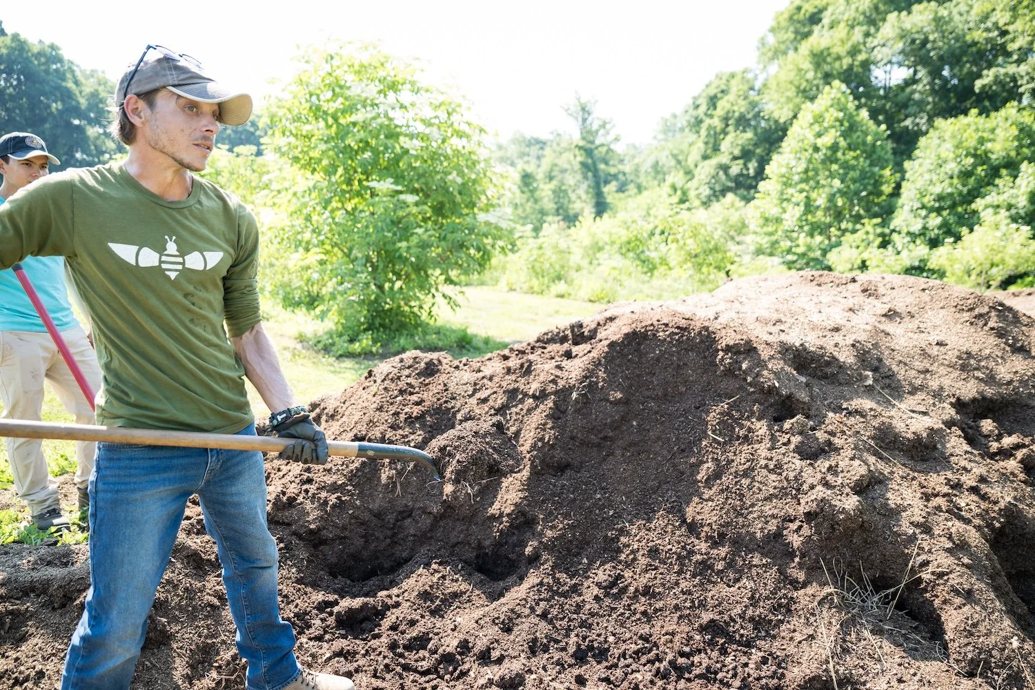 Man in a baseball cap and green long-sleeve t-shirt standing next to a pile of soil holding a shovel