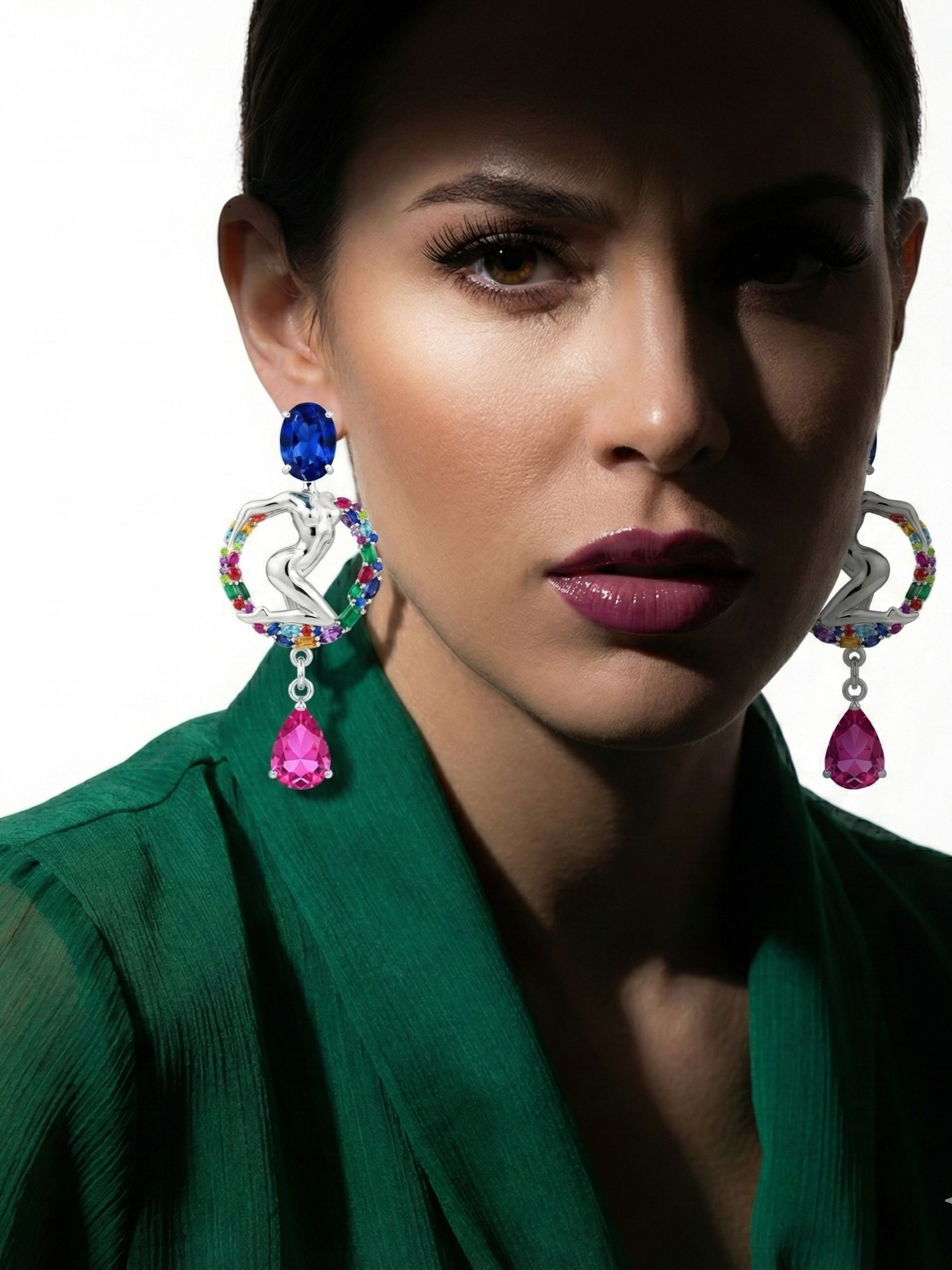 A woman with dark hair, wearing bold makeup and colorful diamond earrings, dressed in a green top, looking directly at the camera.