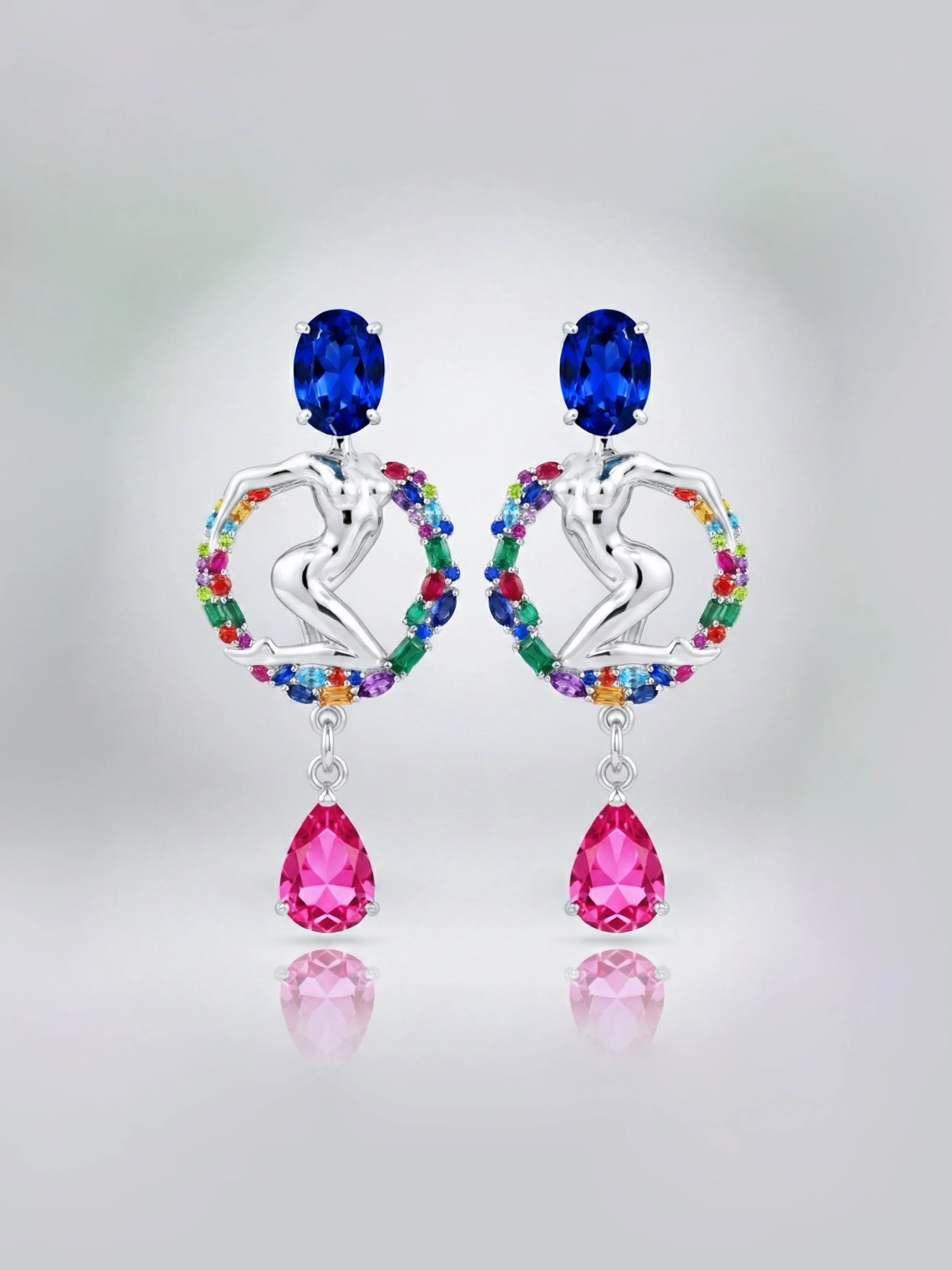 Colorful earrings featuring a human figure silhouette with a blue gemstone head, a rainbow-colored circular frame, and a pink teardrop-shaped gemstone dangling at the bottom.