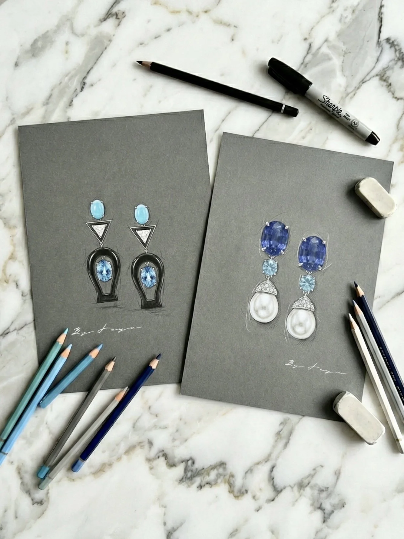 Two sheets of paper on a marble surface with detailed sketches of earrings, surrounded by colored pencils, pens, and erasers.