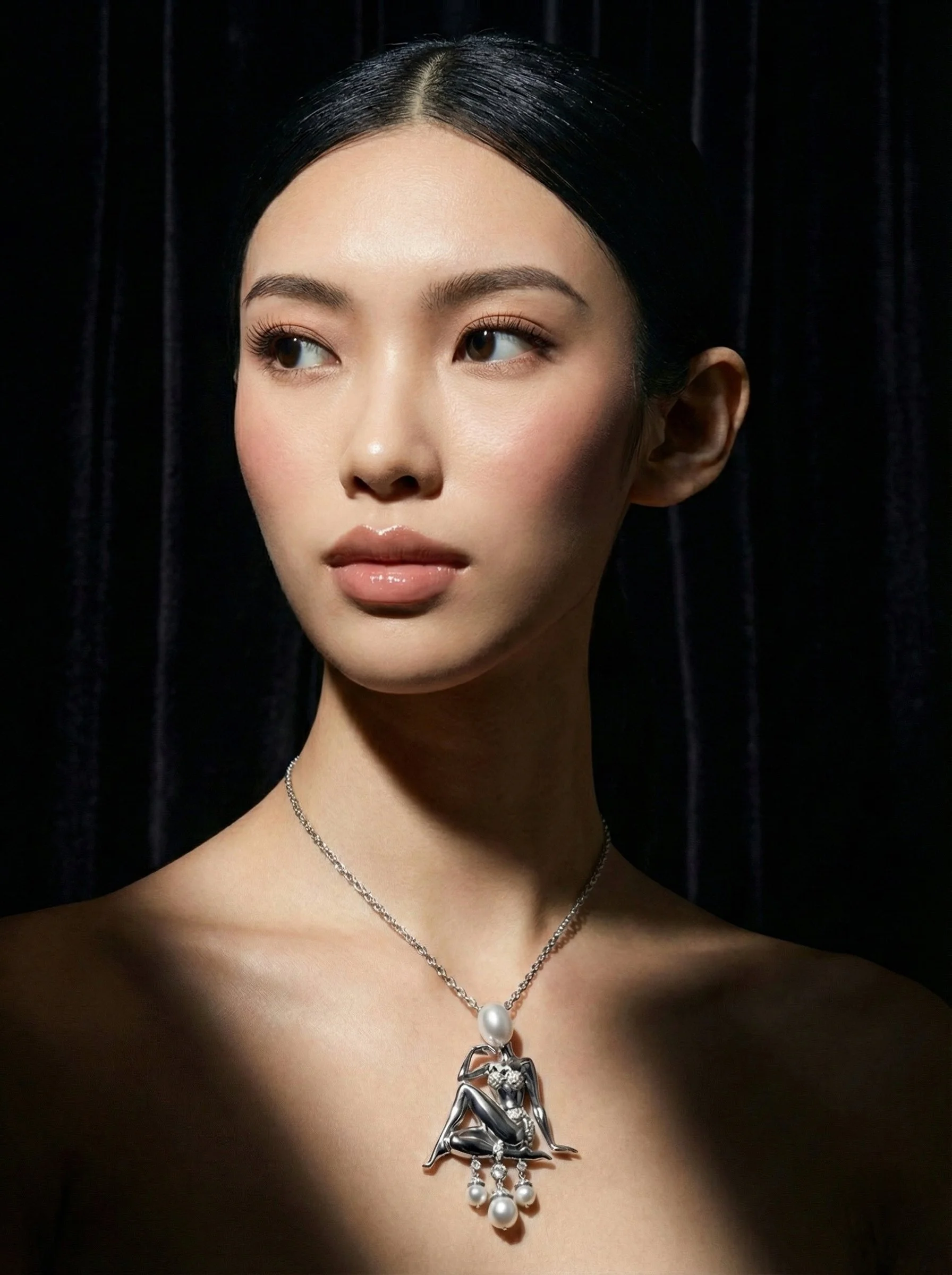 A woman with black hair and a necklace with a large pendant featuring a metallic sculpture of a seated figure with pearls, posing against a dark background.