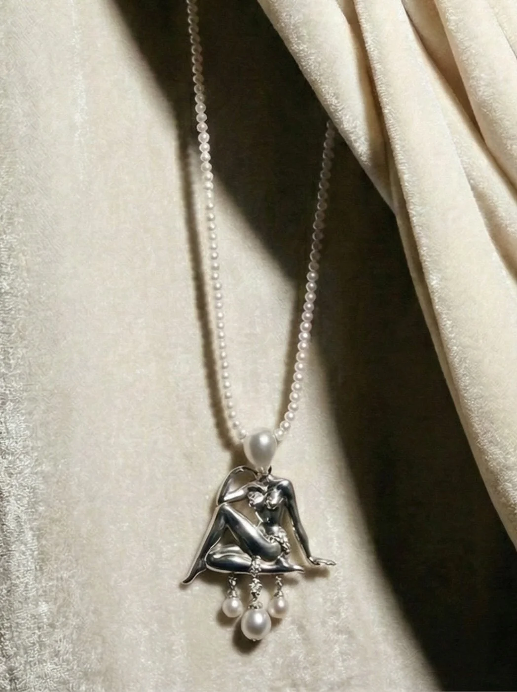 A silver necklace with a pendant of a woman sitting with her knees up and holding her head, decorated with pearls, hanging against beige fabric and a curtain.