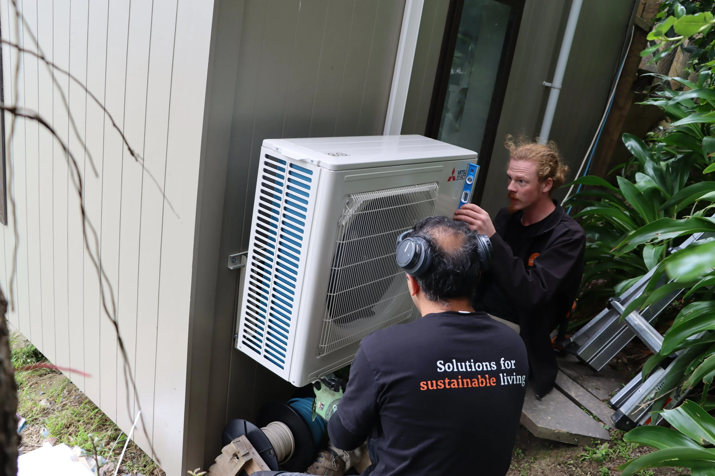 Two Sustainability Trust employees crouched installing outdoor unit of Mistubishi heat pump on exterior wall. One is holding unit onto brackets, one is using spirit level.