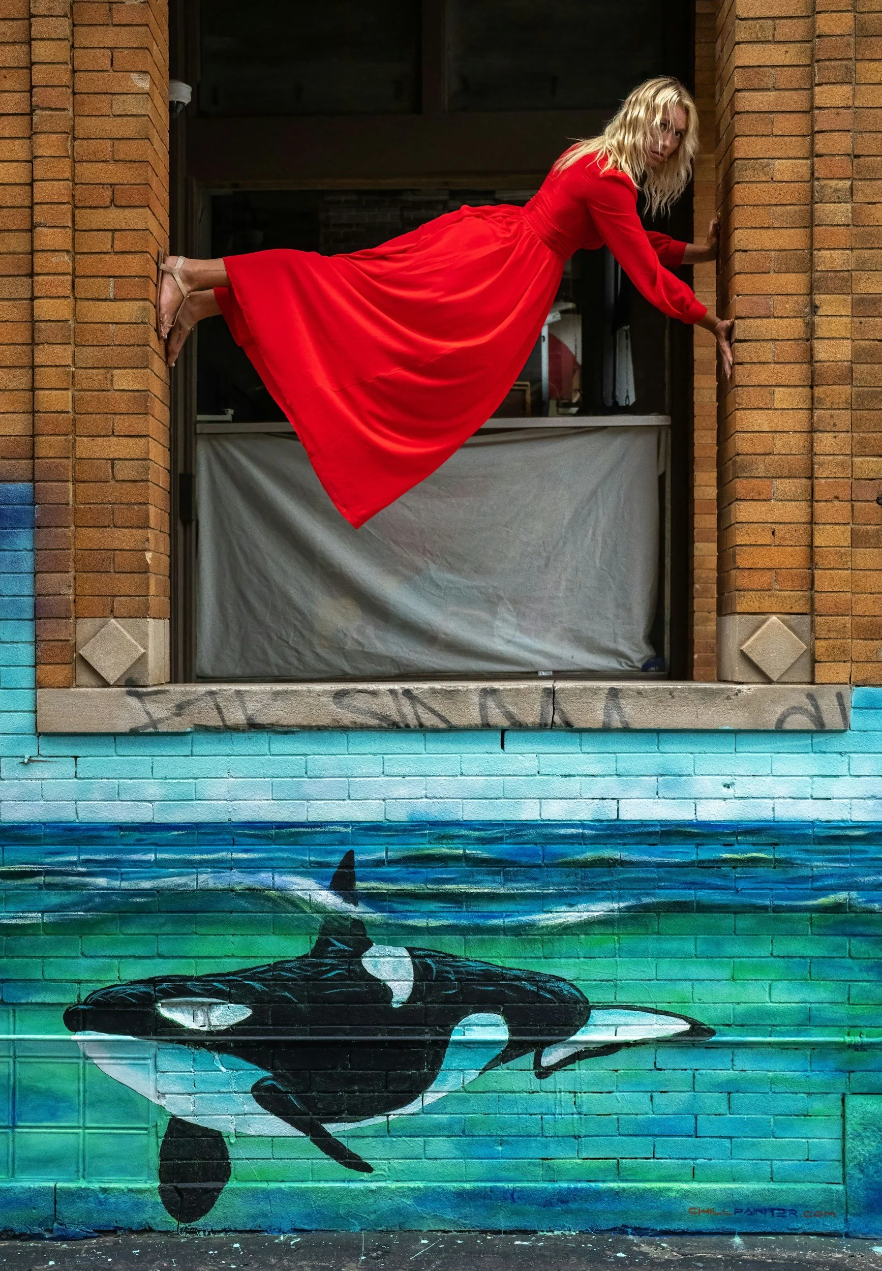 woman in red dress balances her body horizontally within brick wall opening and above a painted mural of a teal sea with orca whales