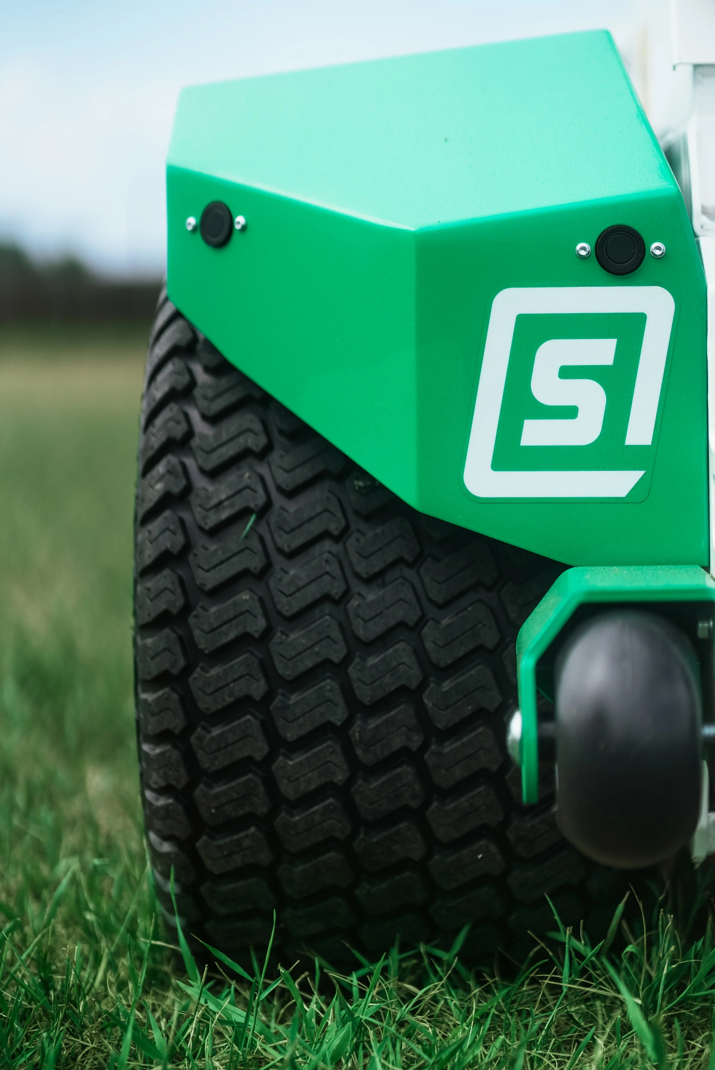 Close-up of a green lawn mower wheel with a textured black tire, green body, and a white logo on the side, on grass.