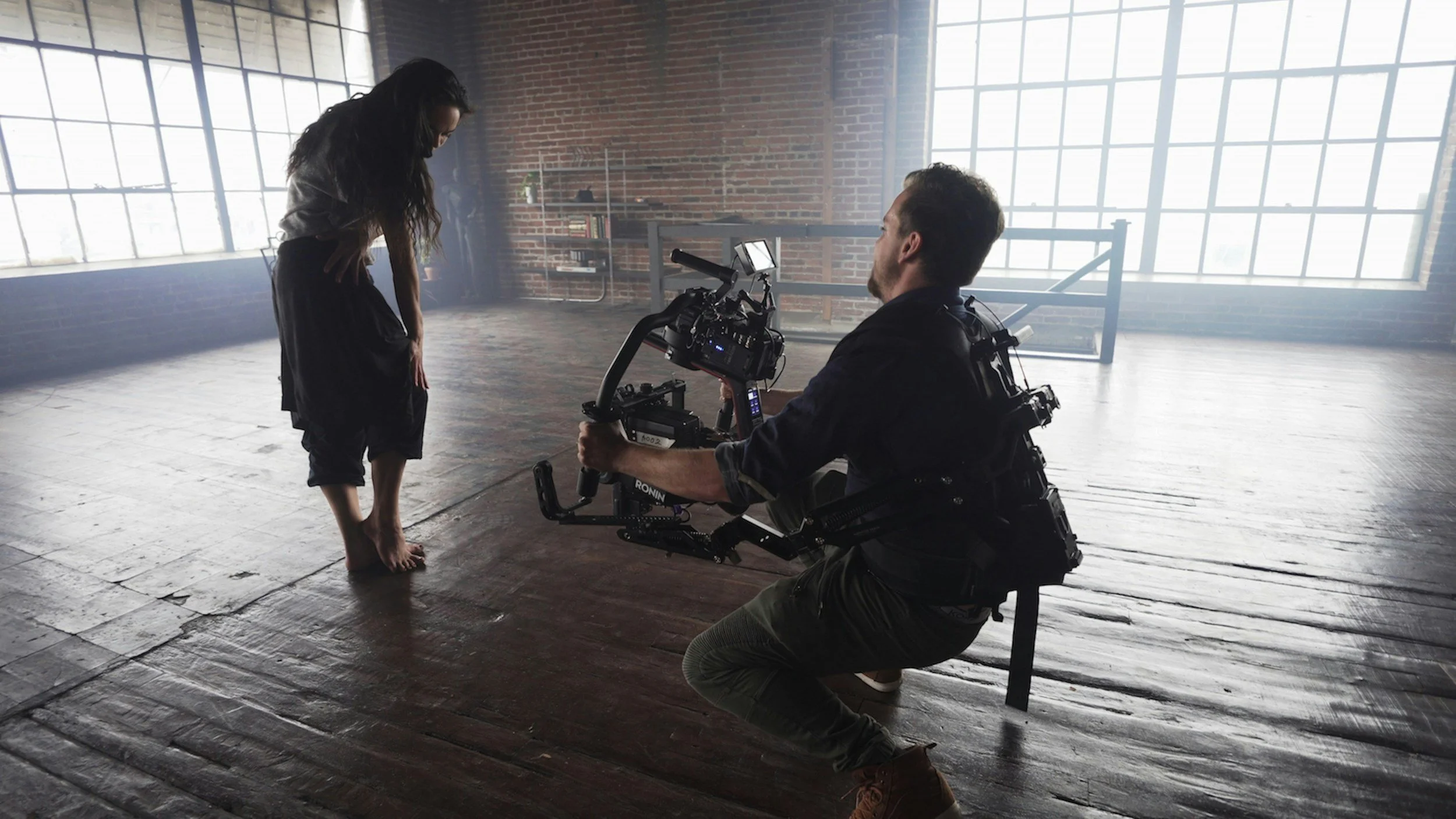 Camera operator filming a performer in a sunlit studio with large windows and a wooden floor