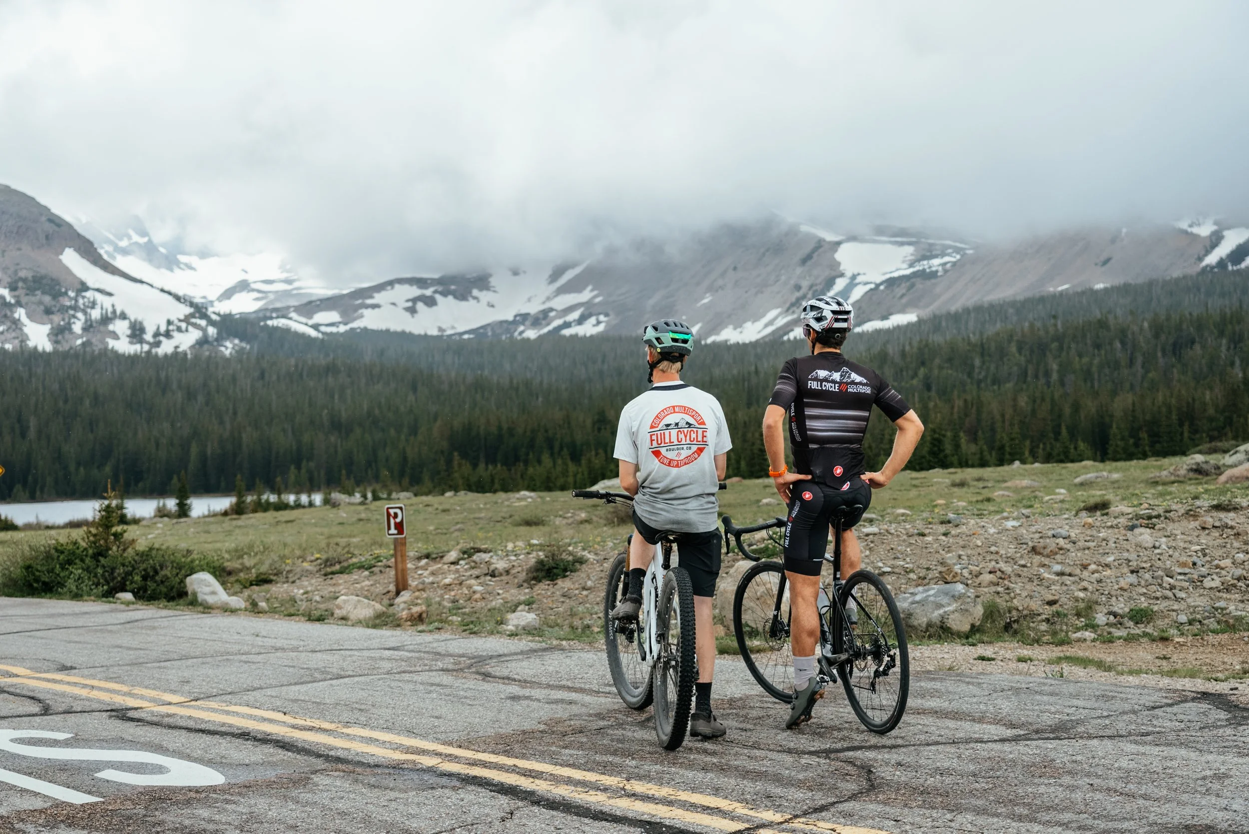 Two cyclists paused beside a mountain road overlooking forest and snowy peaks