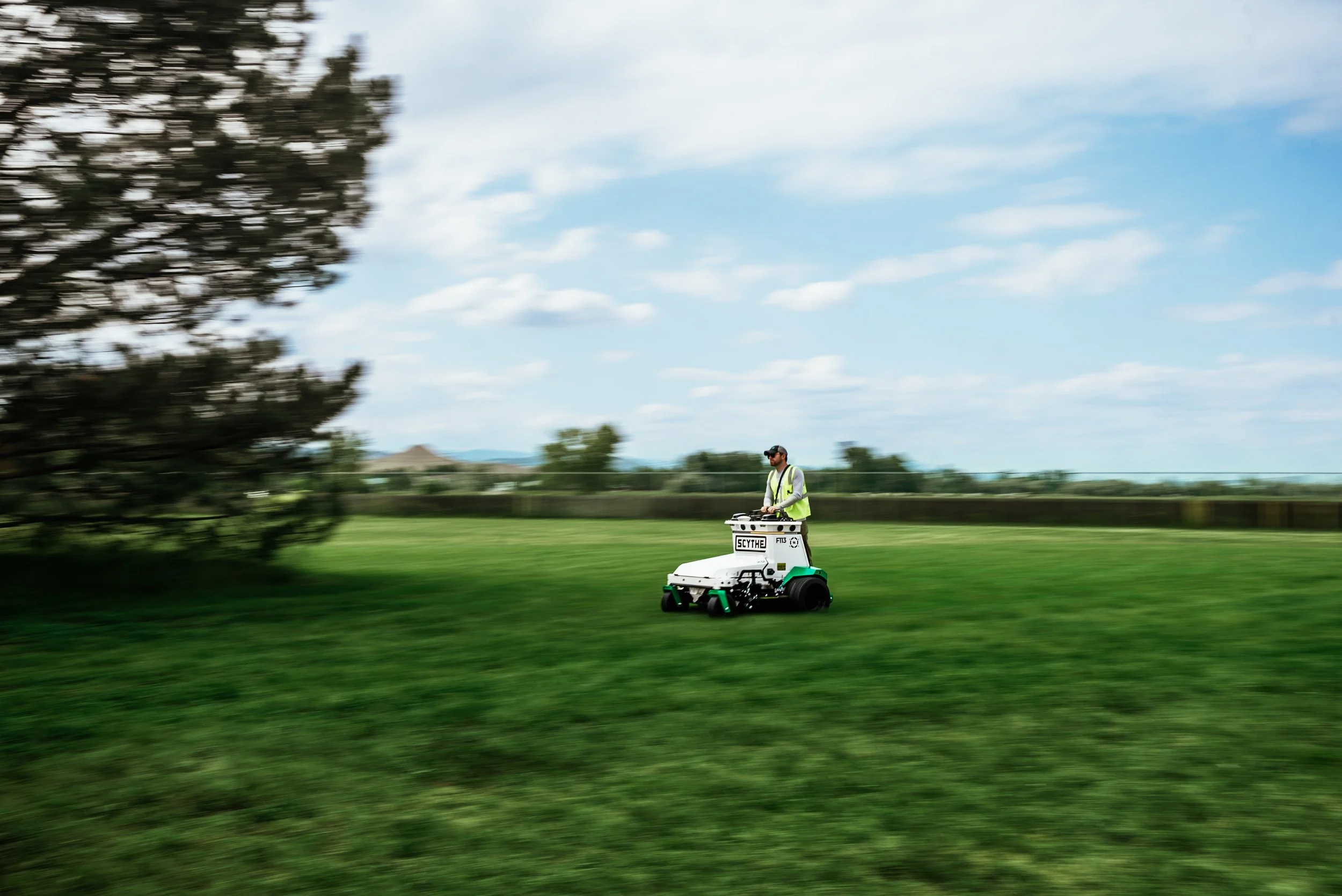 A person operating a robot lawn mower on a large grassy field with trees and a blue sky with clouds in the background.
