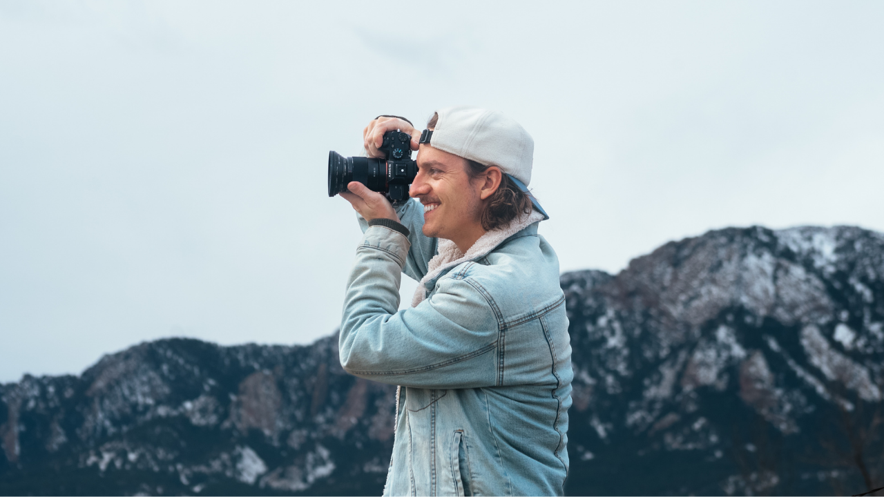 Man in light denim jacket and baseball cap taking a photo with a camera outdoors near mountains