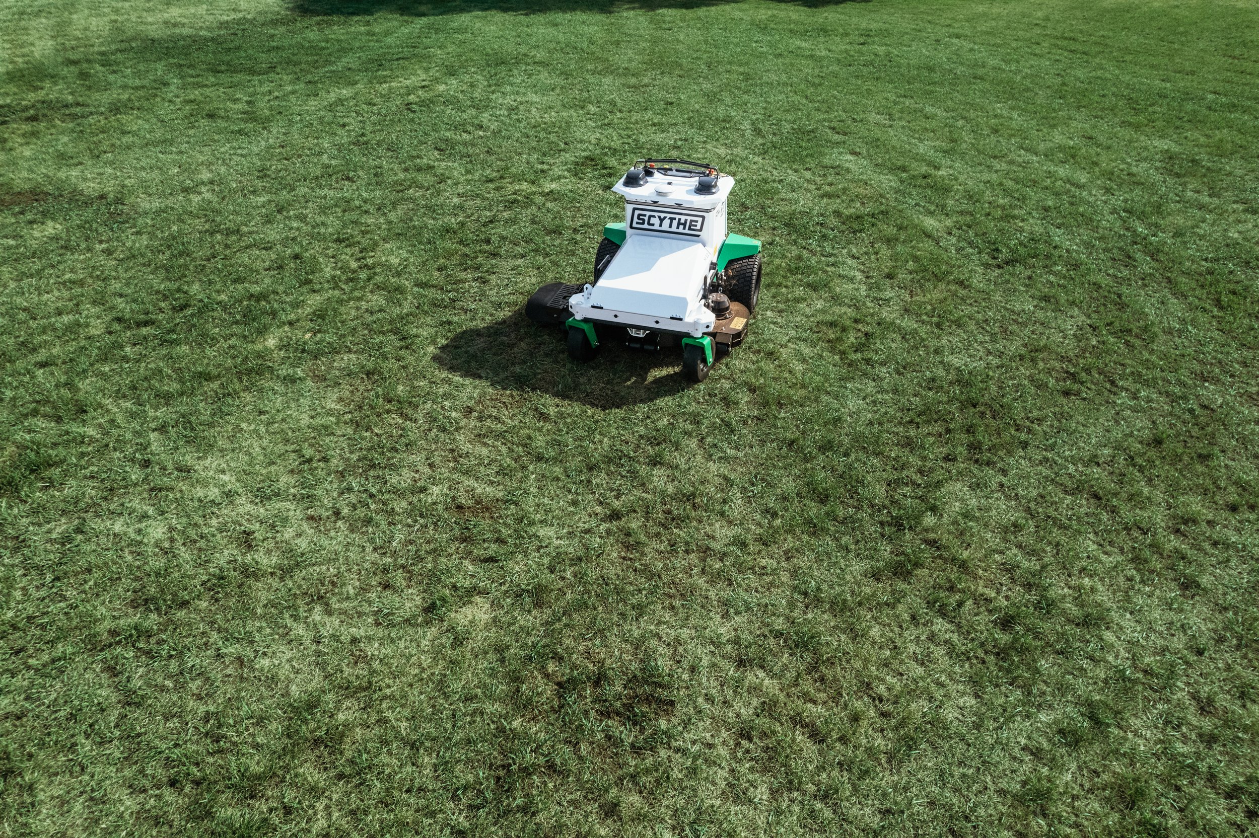 A small robot with a white body and green accents on a grassy lawn, labeled 'SCYTHE' on the front.