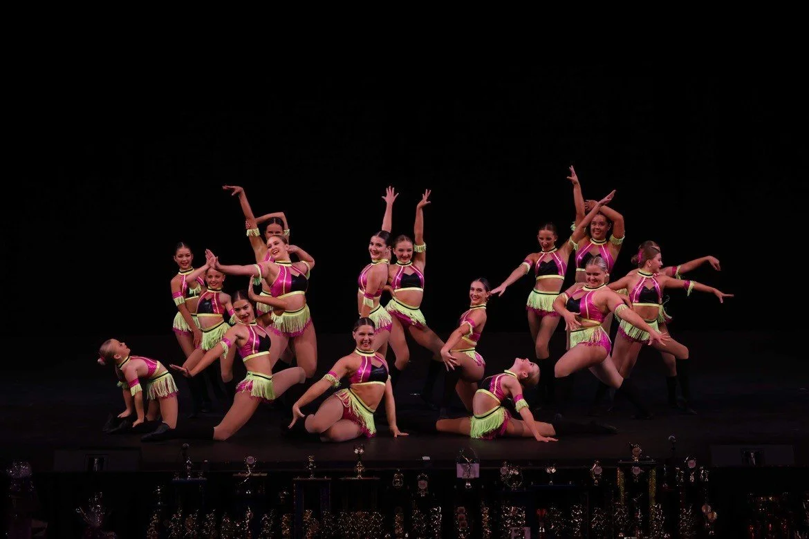 Group of young female dancers in colorful costumes on stage, performing a dance routine.