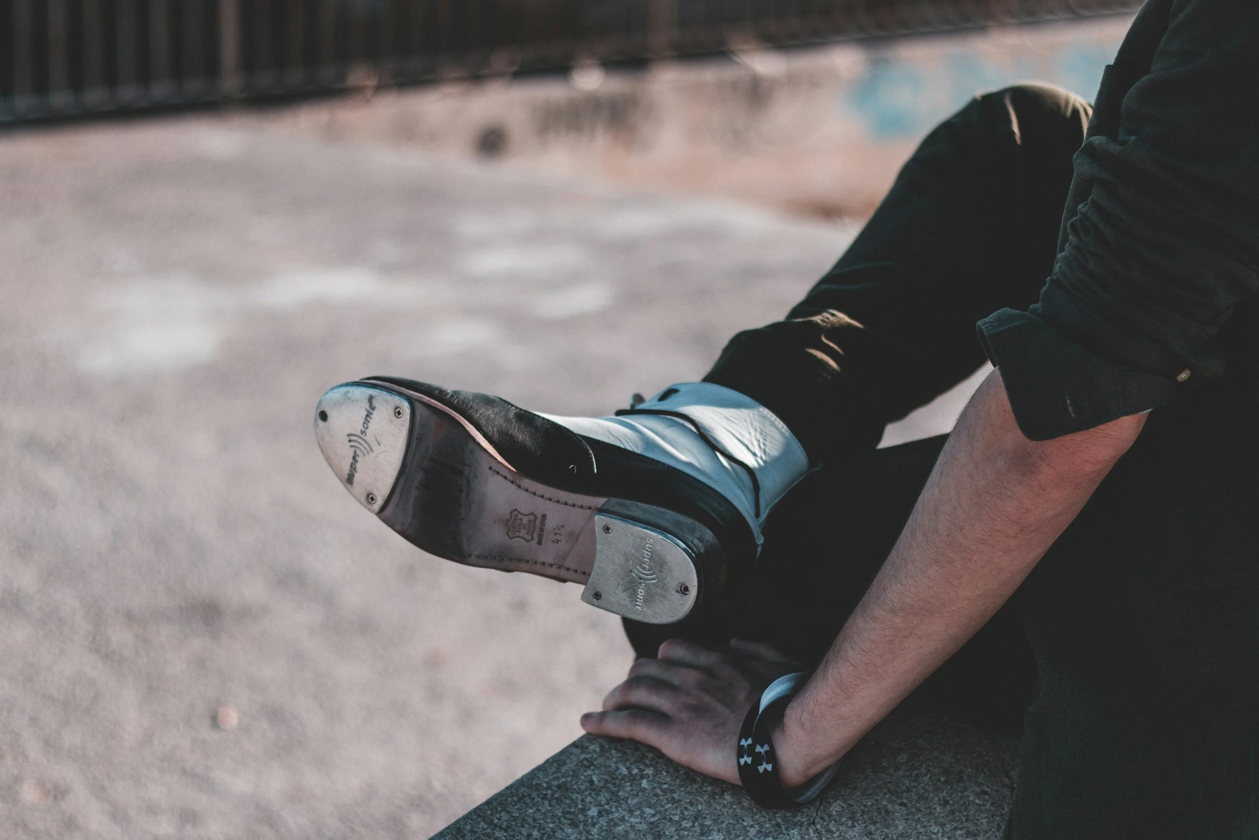 Close-up of a person sitting on a ledge with one leg extended and the foot raised, wearing a white and black ice skate with visible brand markings, in an outdoor skating rink or area.