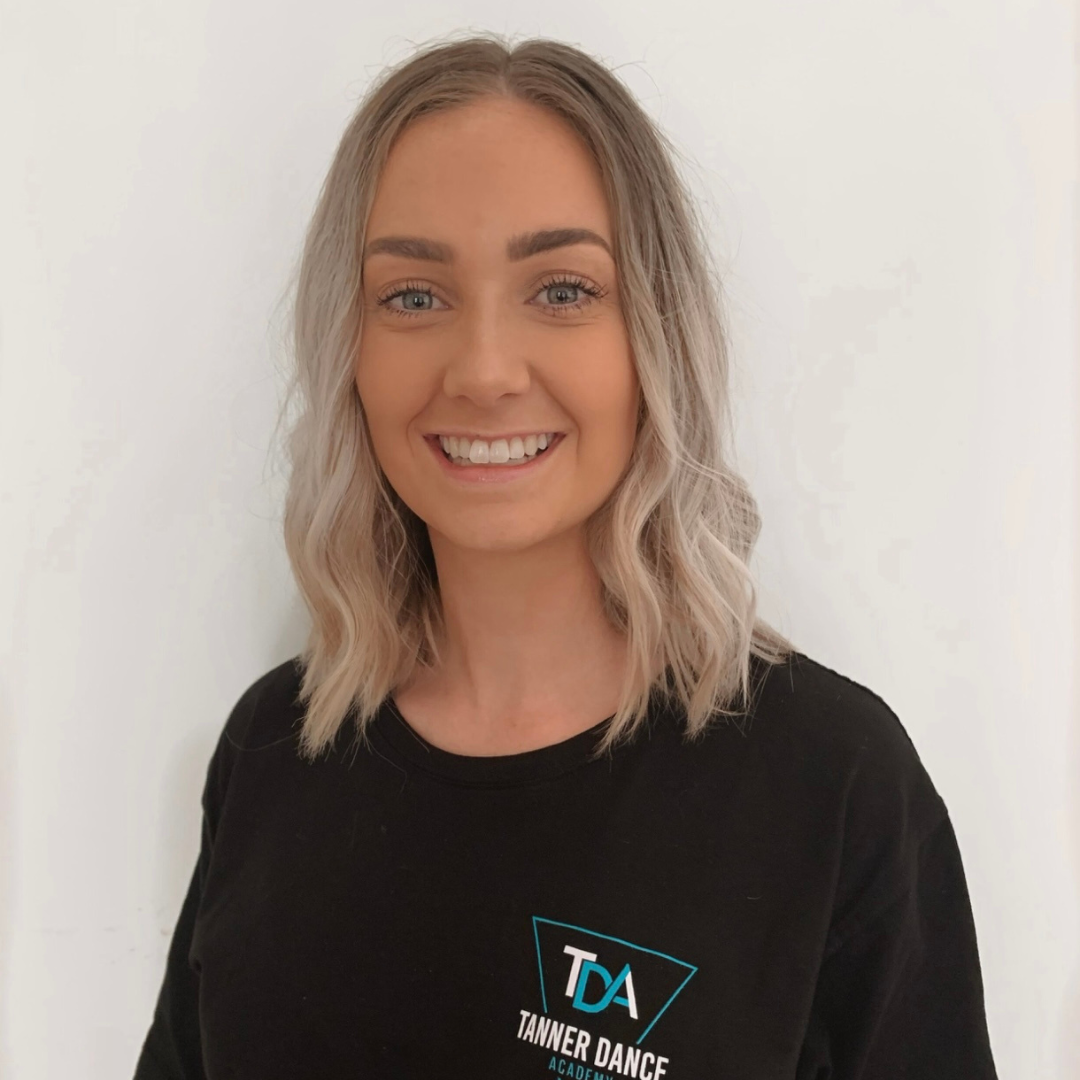 A smiling young woman with wavy blonde hair wearing a black shirt with a Tanner Dance Academy logo, standing against a plain white wall.