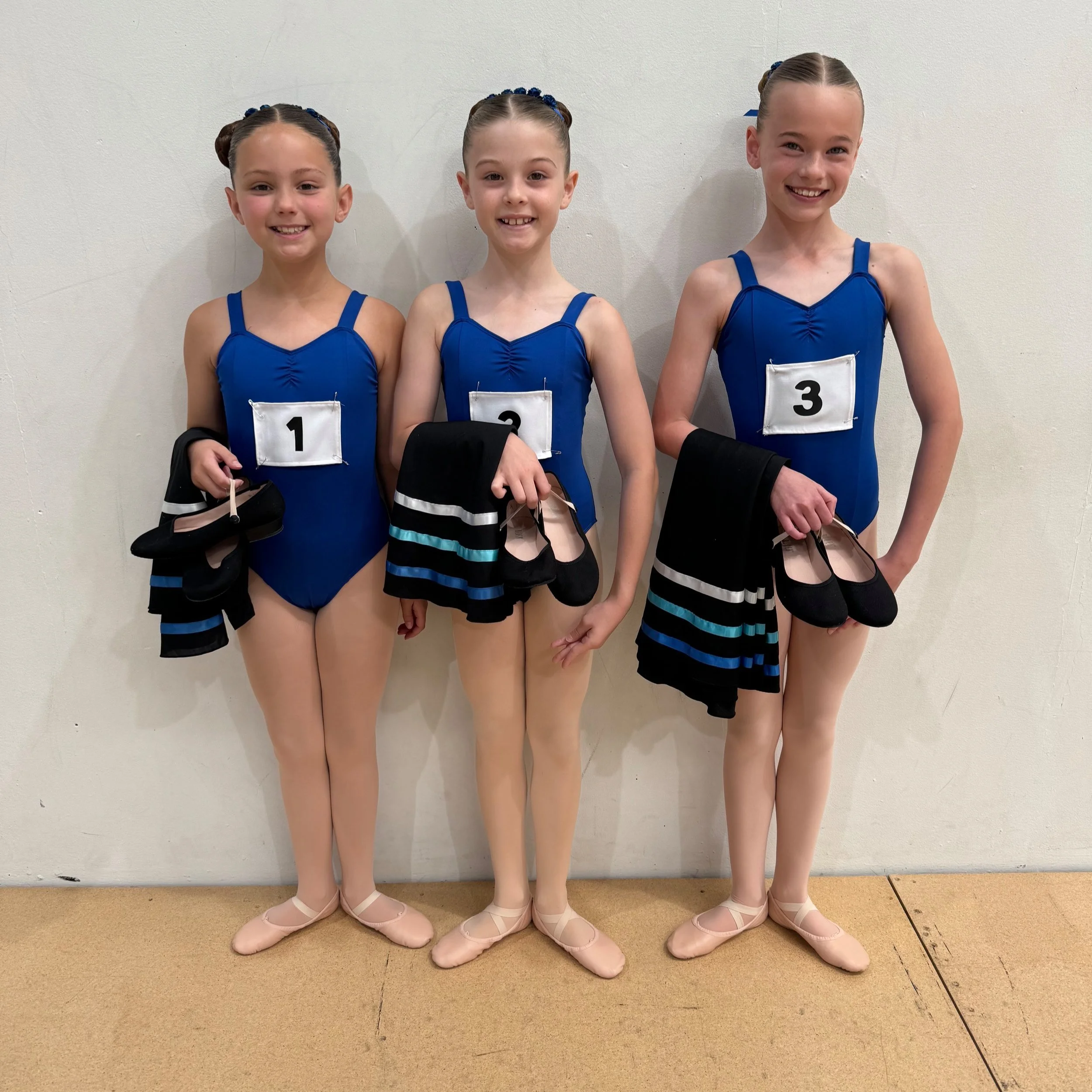 Three young girls in blue leotards with numbers 1, 2, and 3 pinned to their chests, holding black and striped skirts and ballet shoes, standing against a plain wall, smiling.