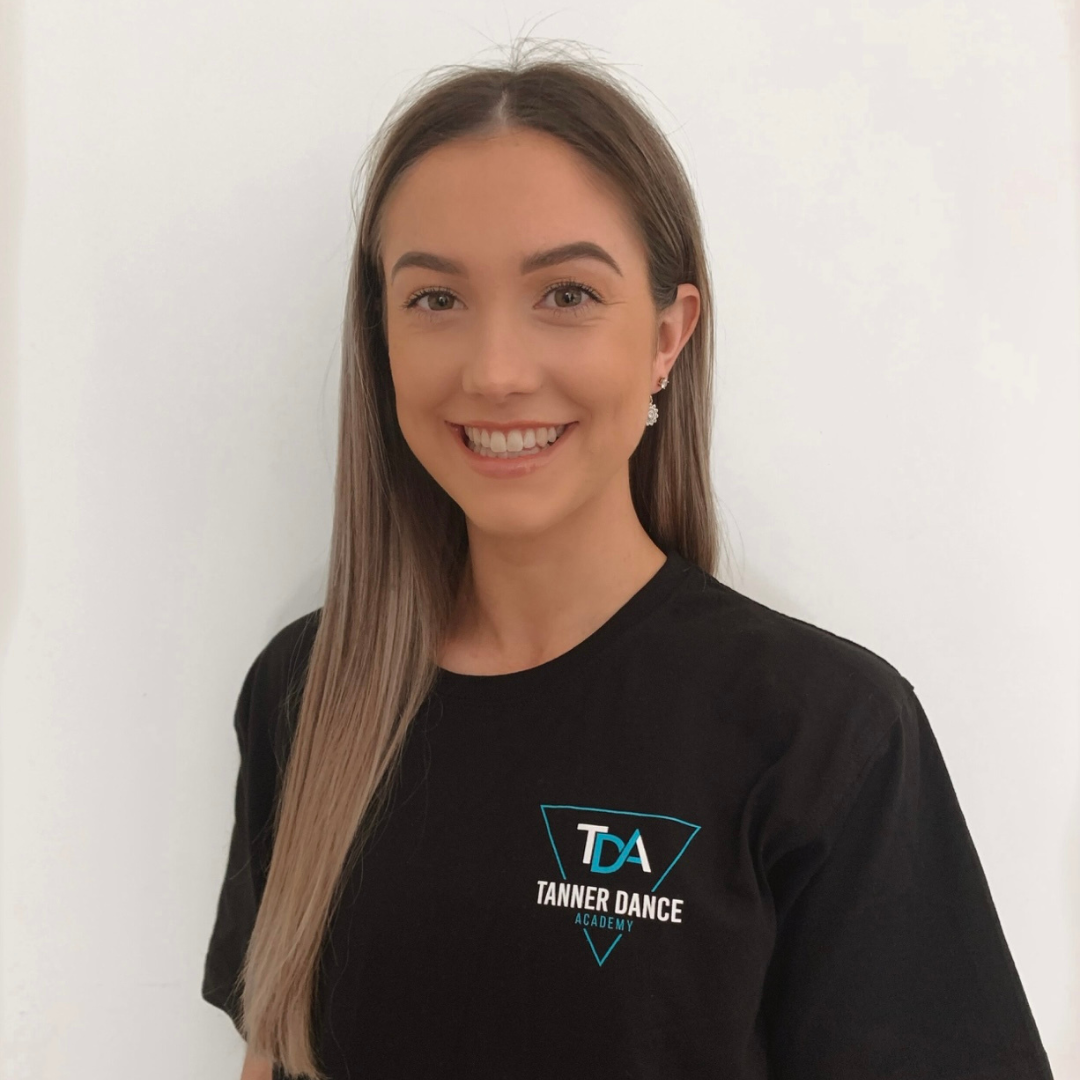 A smiling woman with long brown hair, wearing a black T-shirt with the logo 'Tanner Dance Academy' on it, standing against a plain white background.
