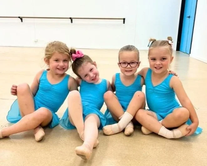 Four young girls in blue leotards sitting on a ballet studio floor, smiling at the camera.