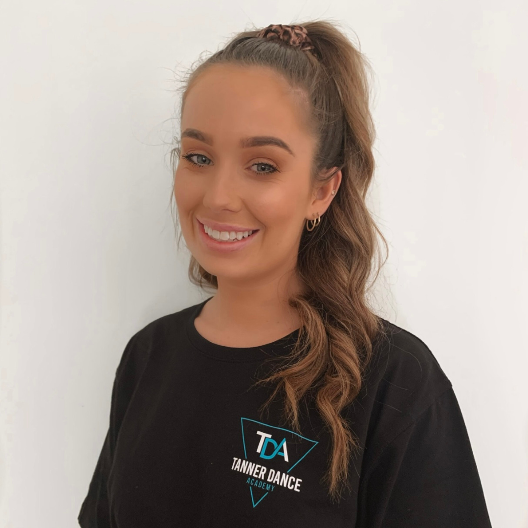 A smiling young woman with long wavy brown hair pinned in a high ponytail with a scrunchie, wearing a black T-shirt with a logo and text for Tanner Dance Academy, standing against a plain white wall.