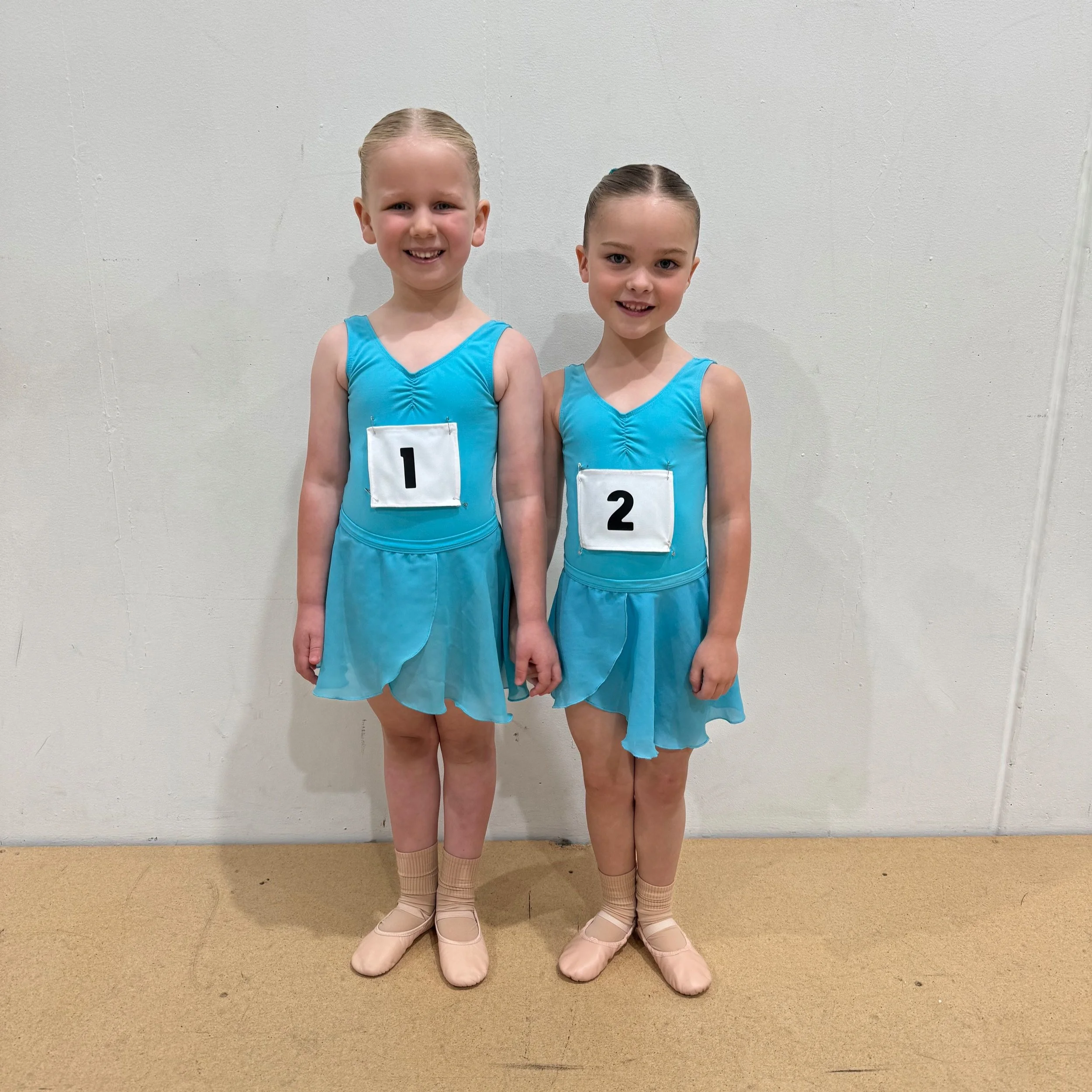 Two young girls in blue dance costumes standing against a plain white wall, each with a number tag (1 and 2), smiling at the camera.