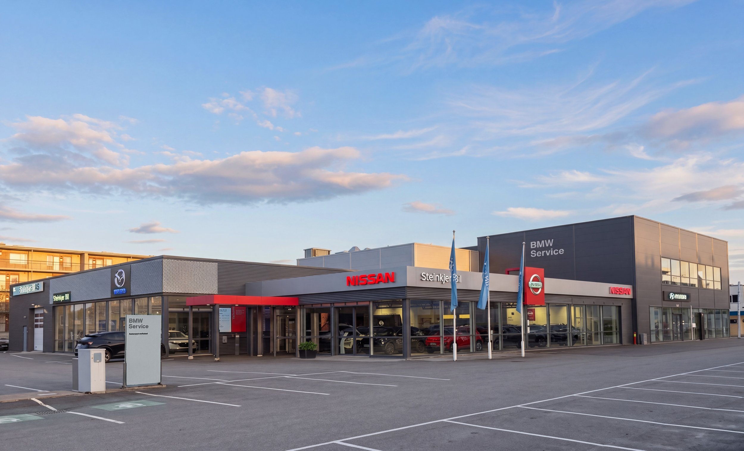 Bilde av Steinkjer Bil på dagtid med synlige Nissan og Mazda skilt, store vinduer som viser biler innendørs og under en lett overskyet himmel.