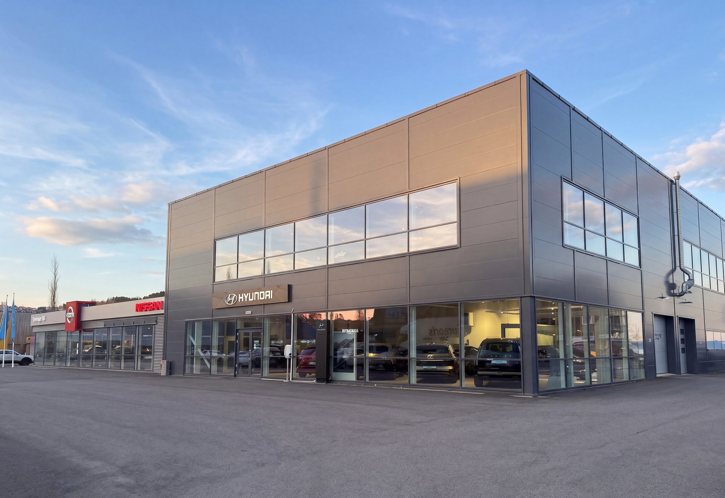 Bilde av Steinkjer Bil på dagtid med synlige Nissan og Hyundai skilt, store vinduer som viser biler innendørs og under en lett overskyet himmel.