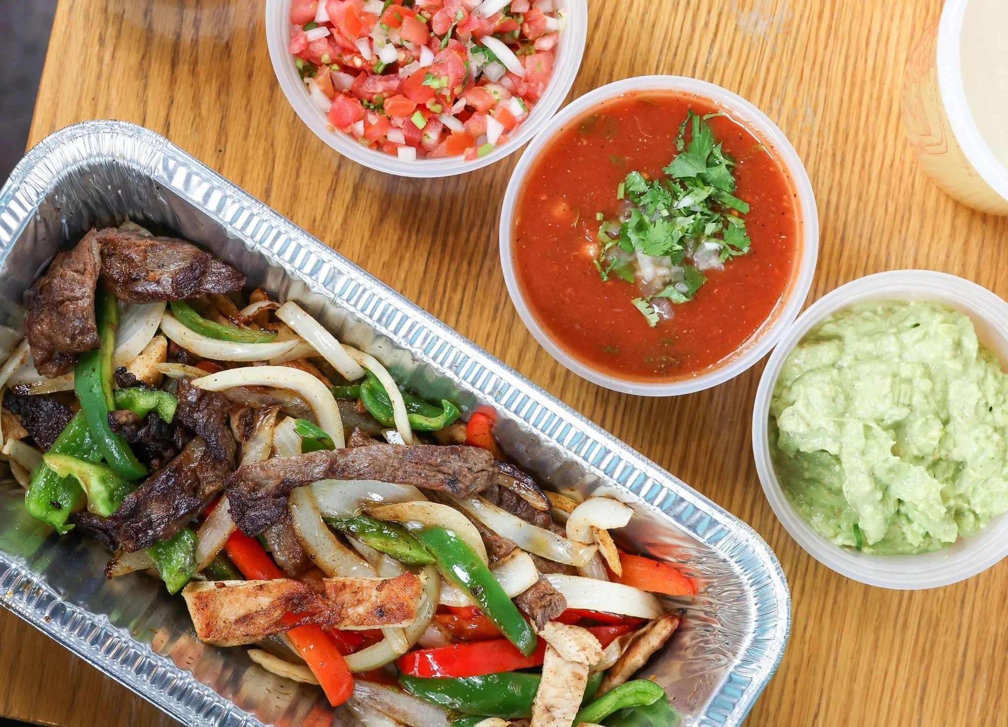 Takeout meal with grilled beef and vegetable stir-fry, bowls of pico de gallo salsa, red sauce, guacamole, and a container of shredded cheese on a wooden table.