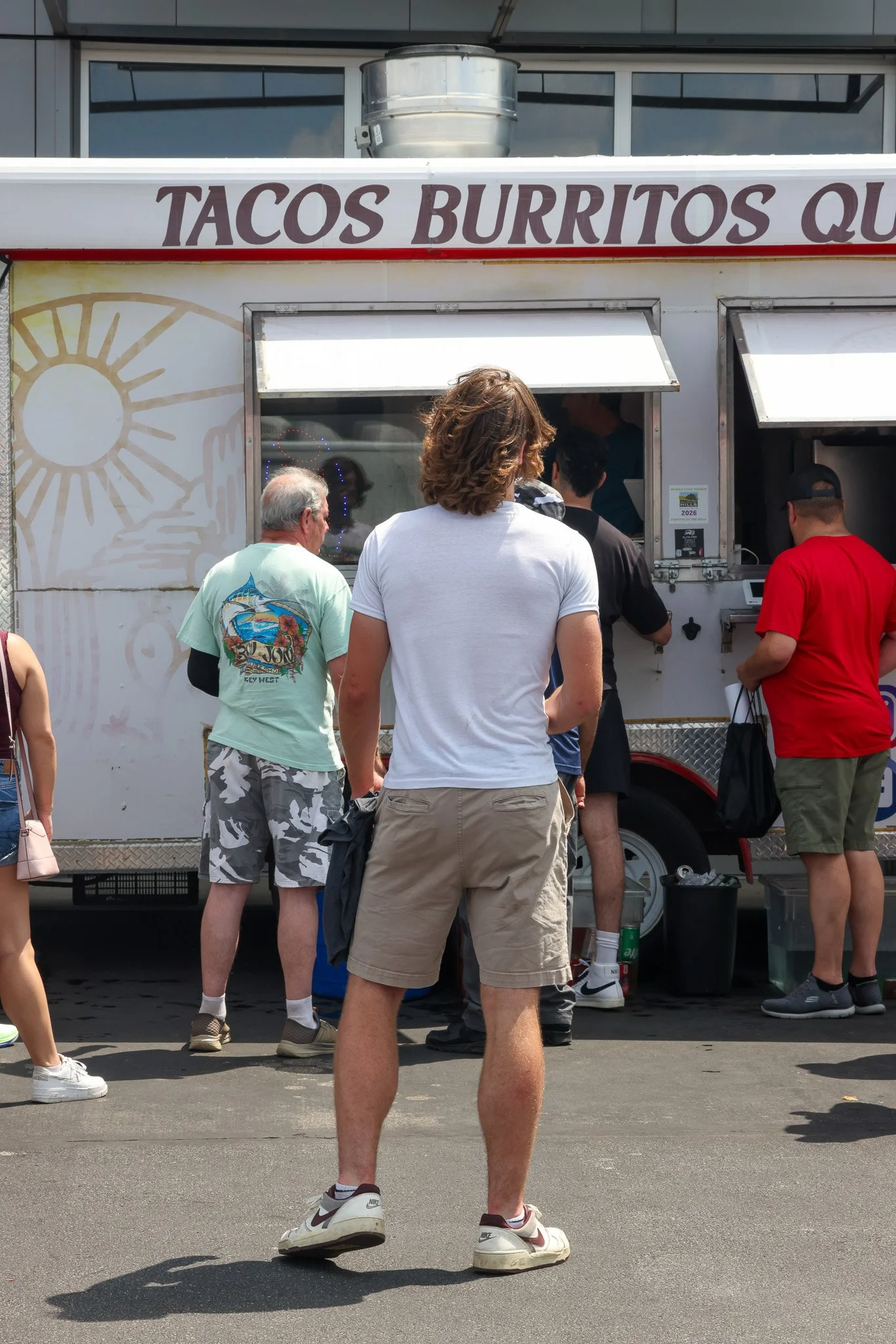 People waiting in line at a food truck called Tacos Burritos Qu., with a man in shorts and Nike sneakers in the foreground.