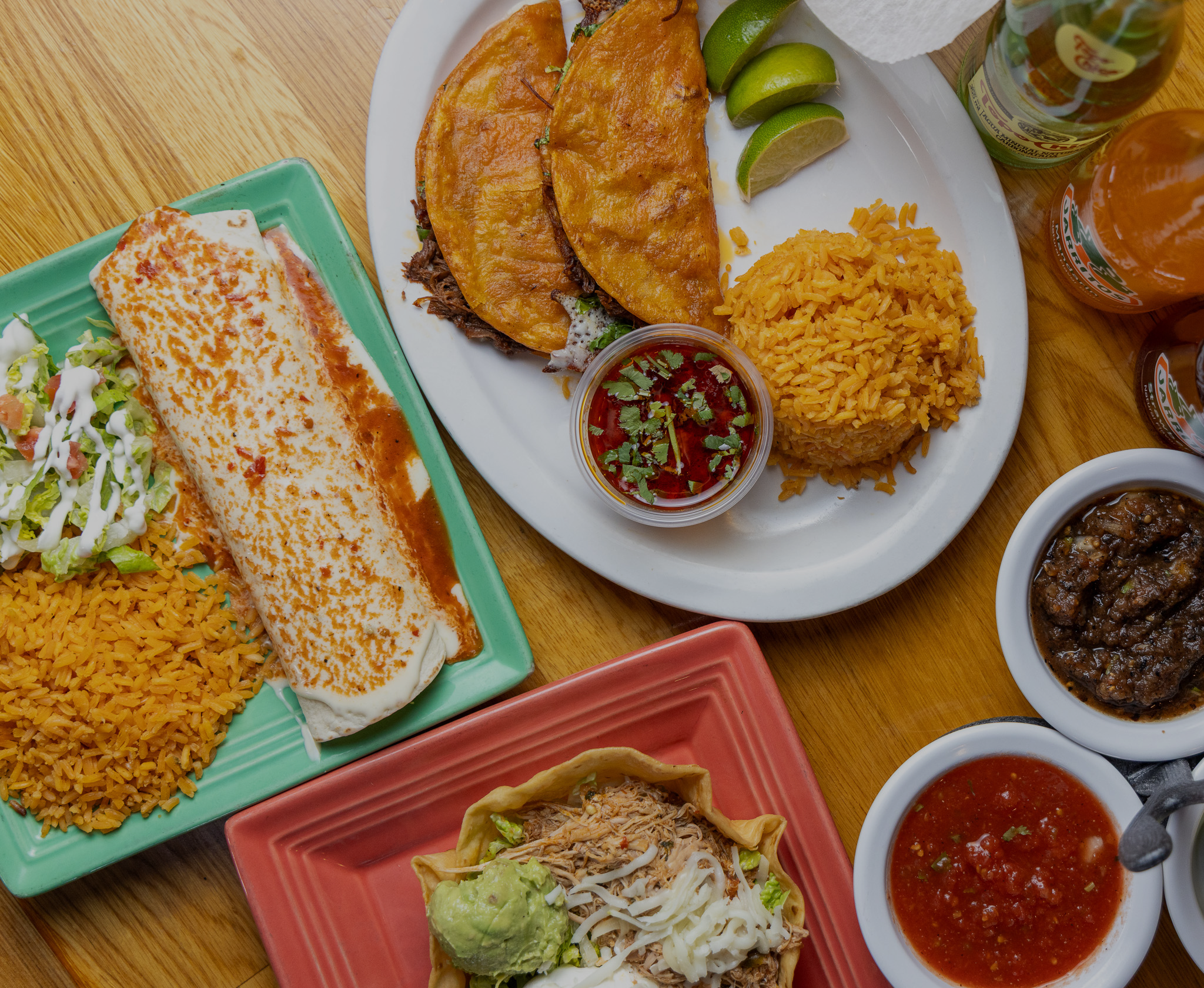 Assorted Mexican dishes on colorful plates including tacos with shredded beef, rice, nachos, salsa, guacamole, salad, and various bottled beverages on a wooden table.