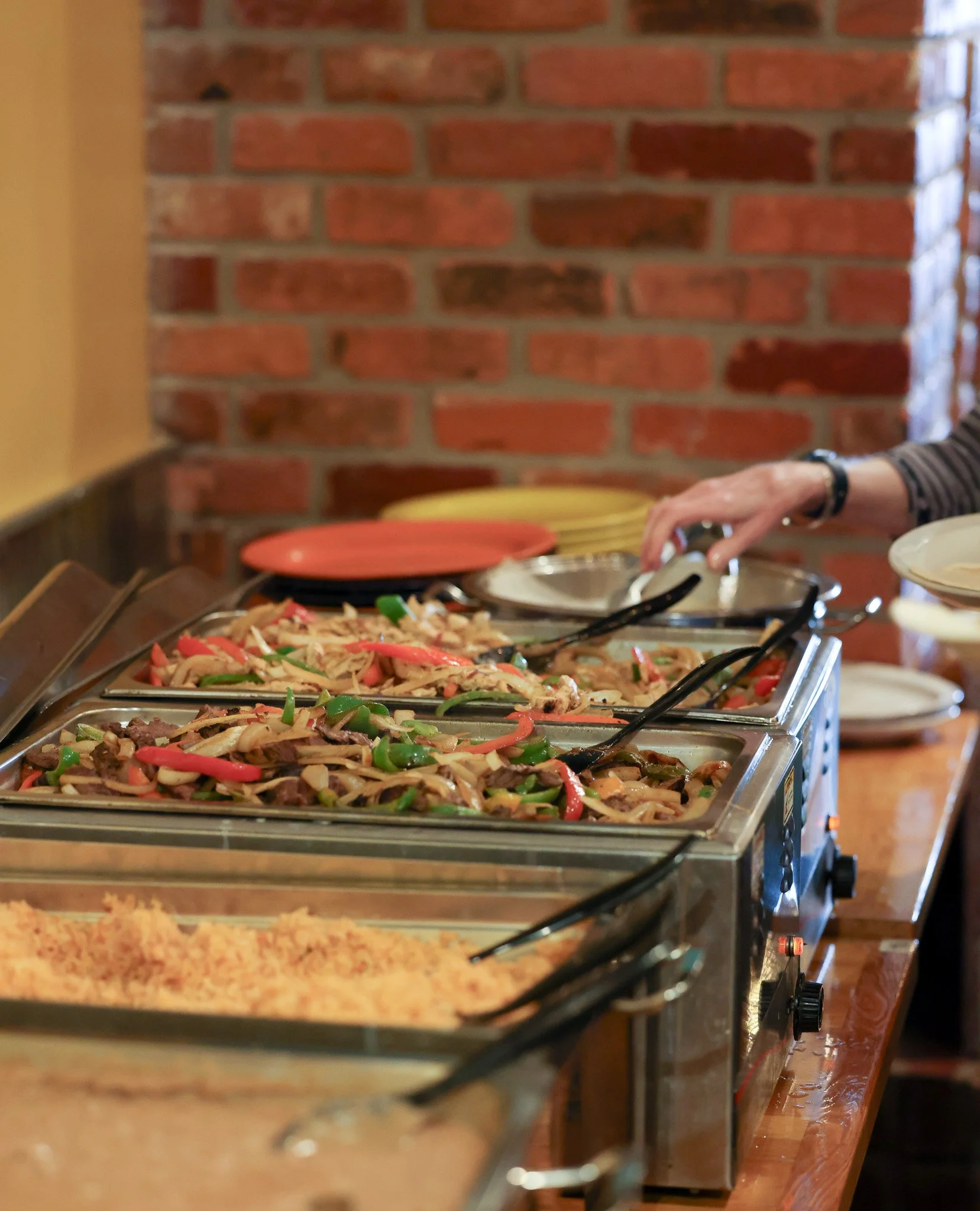 Buffet line with trays of stir-fried vegetables and meats, dishes with peppers, onions, and other vegetables, in a restaurant setting with a brick wall background and plates stacking on the side.