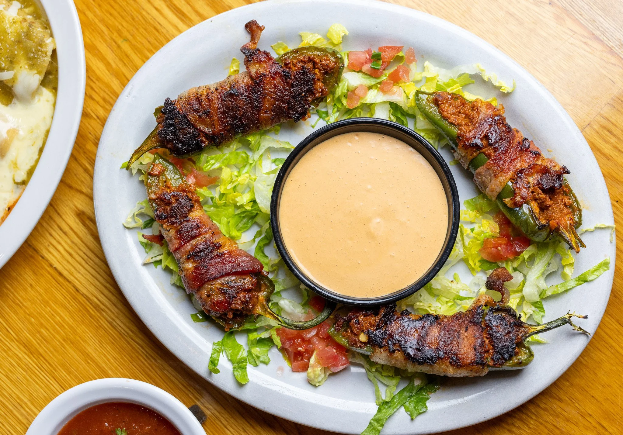 Grilled bacon-wrapped jalapeño poppers on a bed of lettuce with chopped tomatoes, accompanied by a bowl of creamy dipping sauce and a small dish of salsa, on a wooden table.