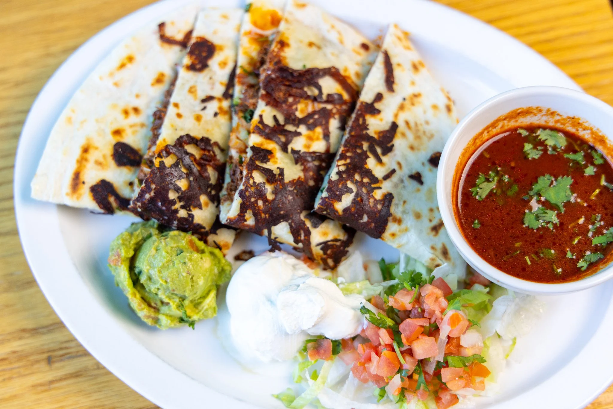 A white plate of Mexican food including tacos with various fillings, a bowl of red sauce garnished with cilantro, guacamole, and a side salad with diced tomatoes and vegetables.