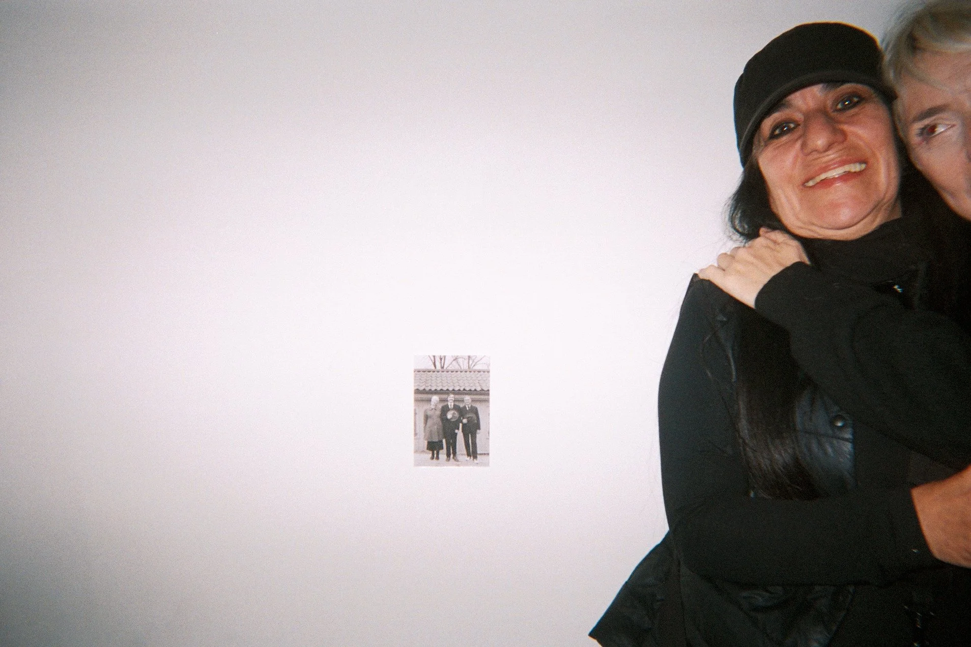 Partially visible woman wearing a black hat and black jacket, smiling and hugging someone, with a black-and-white photo on the white wall behind her.