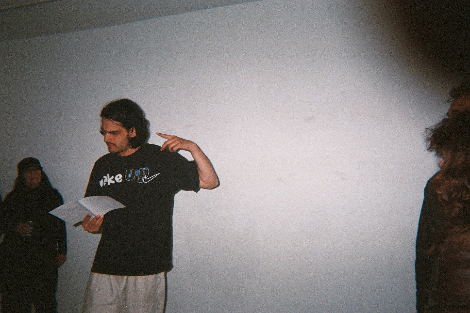 A man with shoulder-length dark hair and mustache reading a book, wearing a black Nike t-shirt, in an indoor setting with others around him.