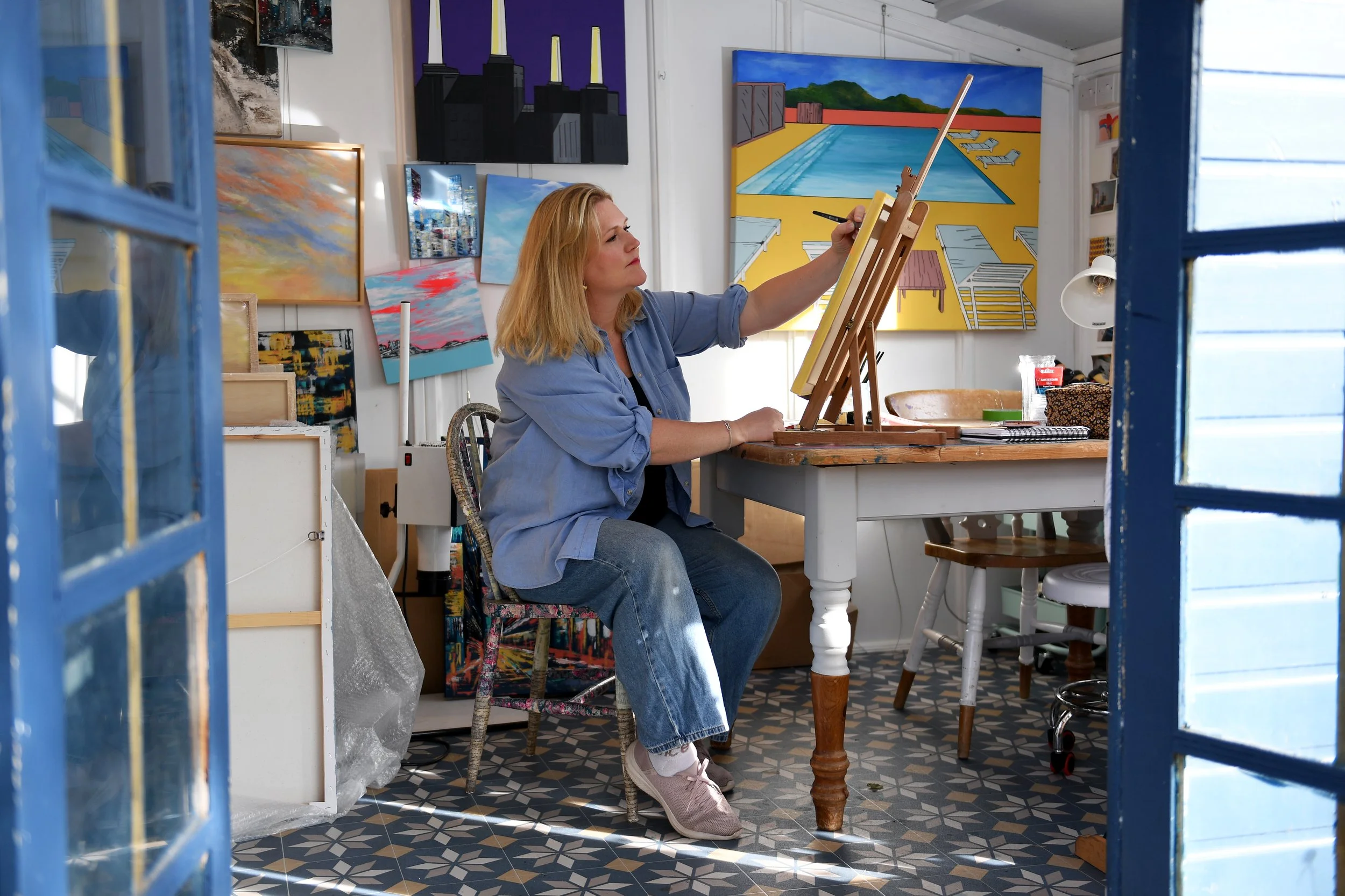 Seated artist painting with paintings on wall behind them
