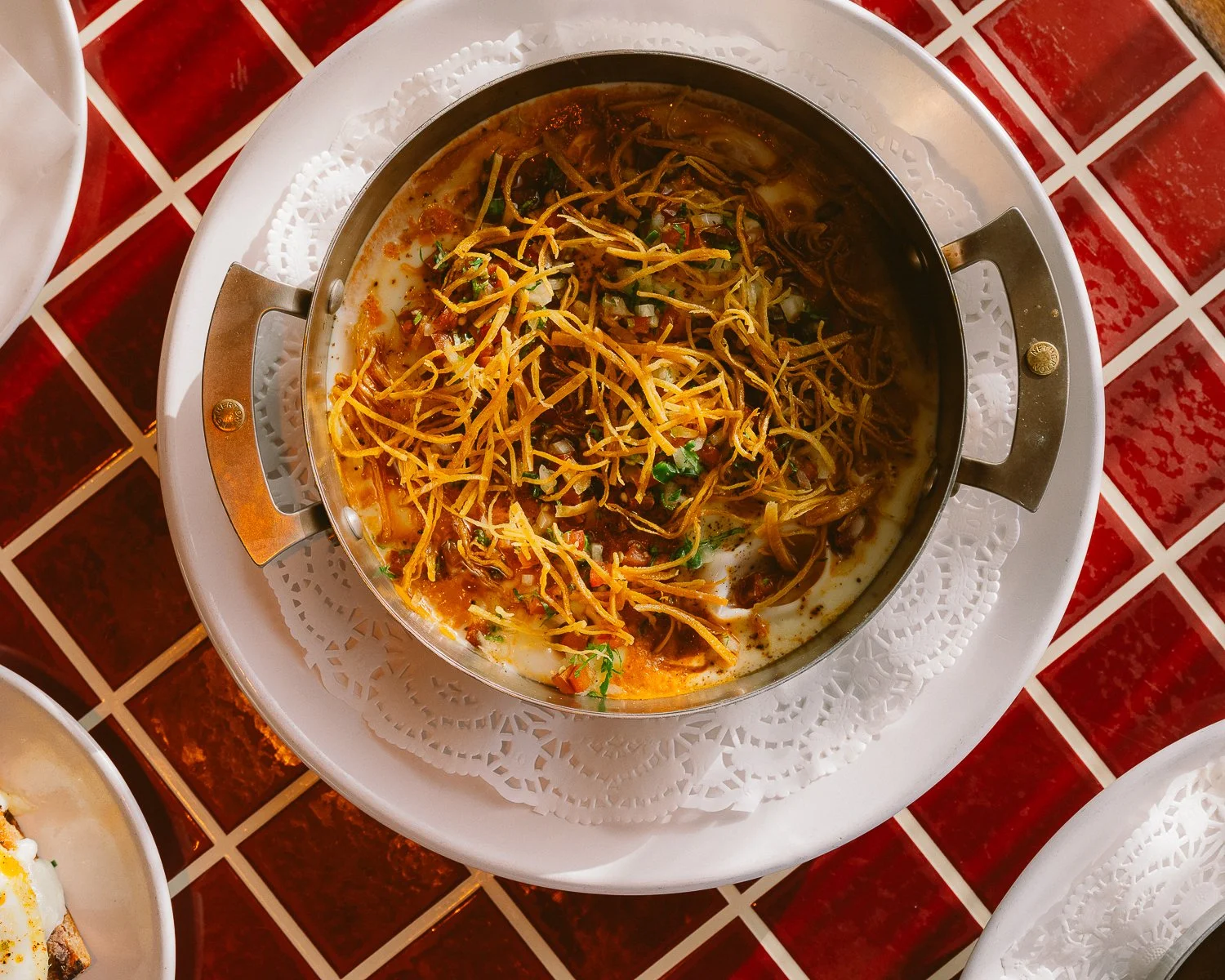 A bowl of Mexican shredded beef and rice topped with shredded cheese, chopped onions, and cilantro, served on a white doily and placed on a white plate, on a red tiled table.