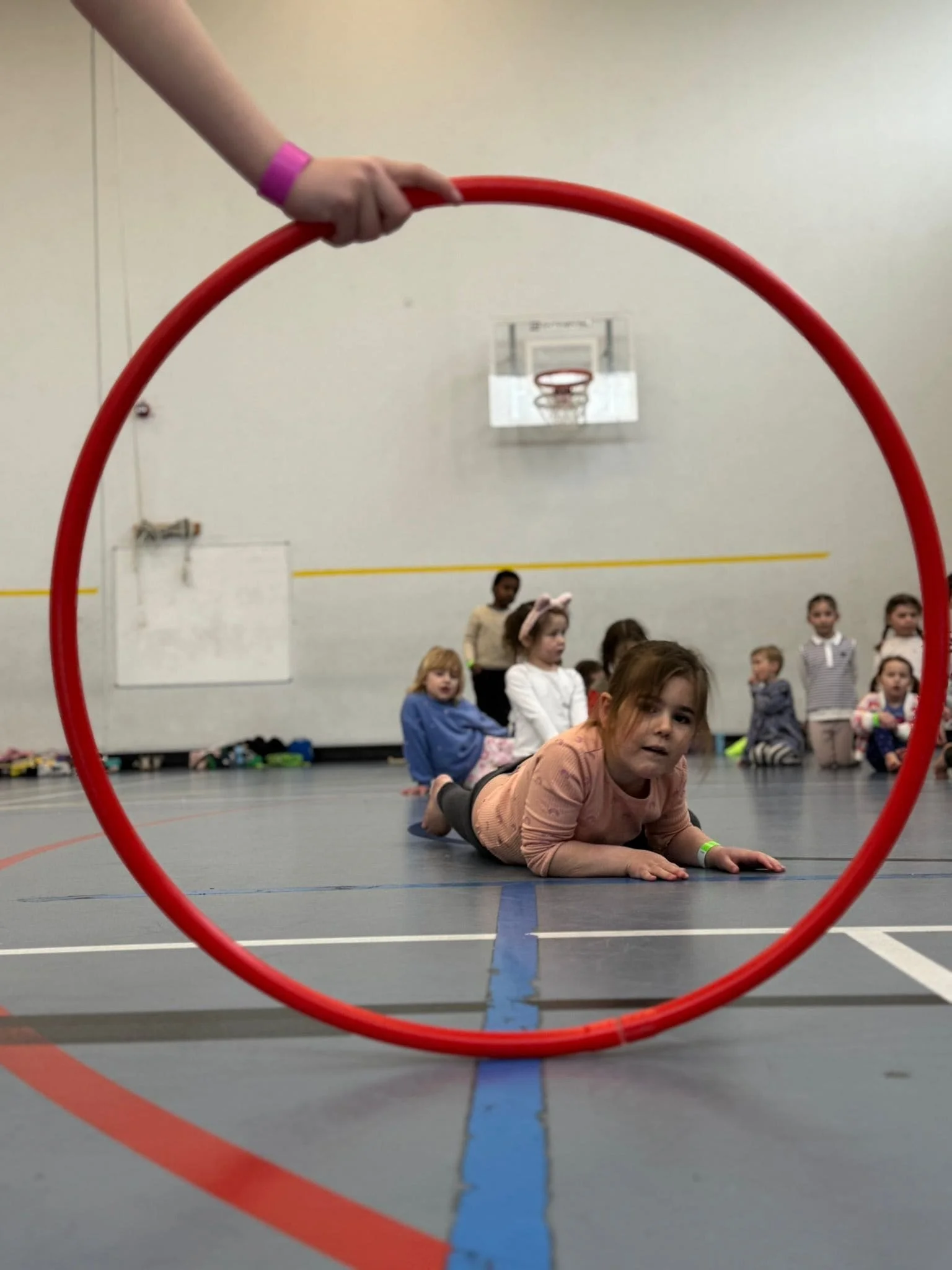 Seen through a red hoop held up in the foreground, a girl crawls across the sports hall floor while other children wait their turn by the wall.