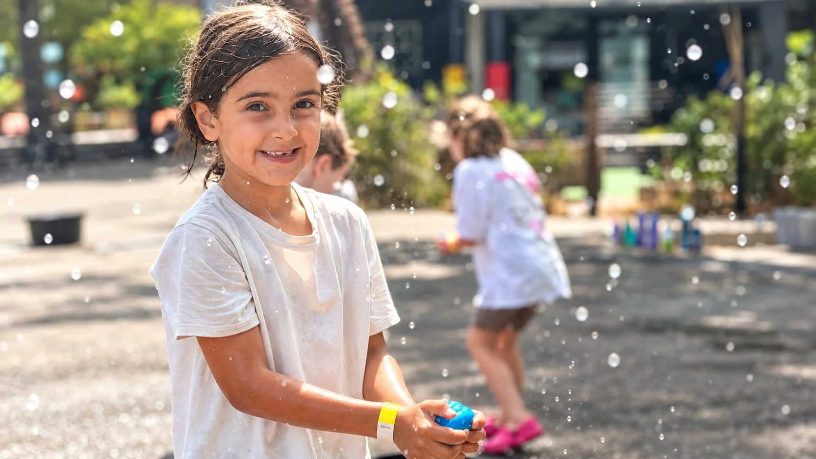 A smiling girl holding a water balloon during outdoor games; an edited GymLab Camps photograph.