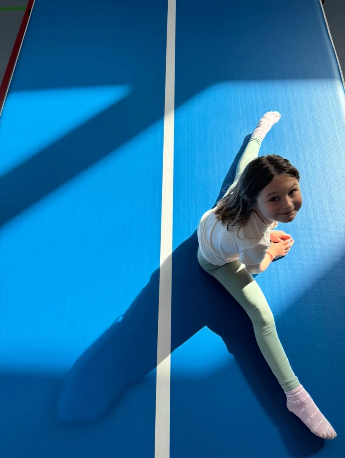 Seen from above, a girl in a split on a blue air track smiles up at the camera.