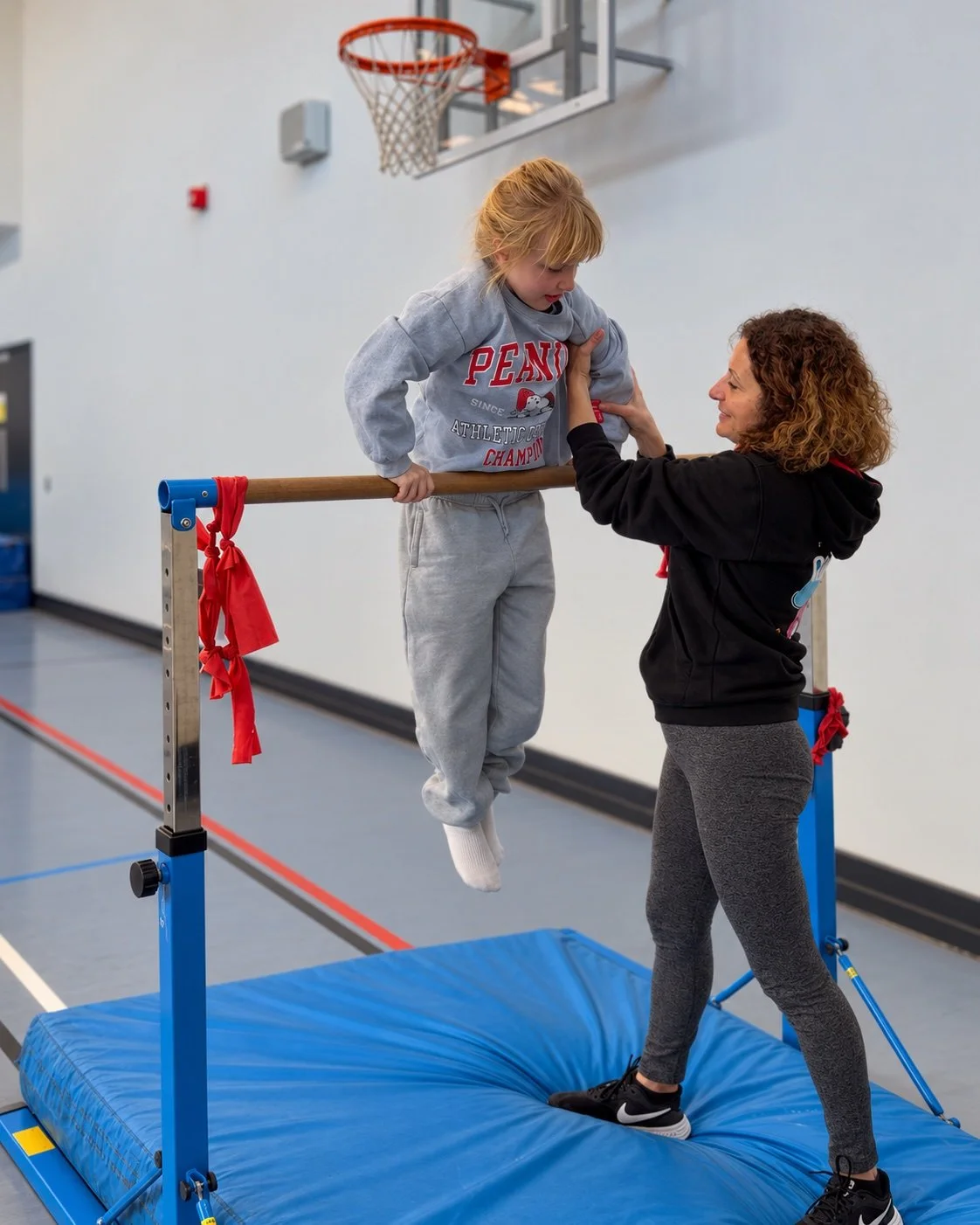 A boy holds himself up on a bar while a coach stands in front of him with her hands ready; an edited GymLab photograph.