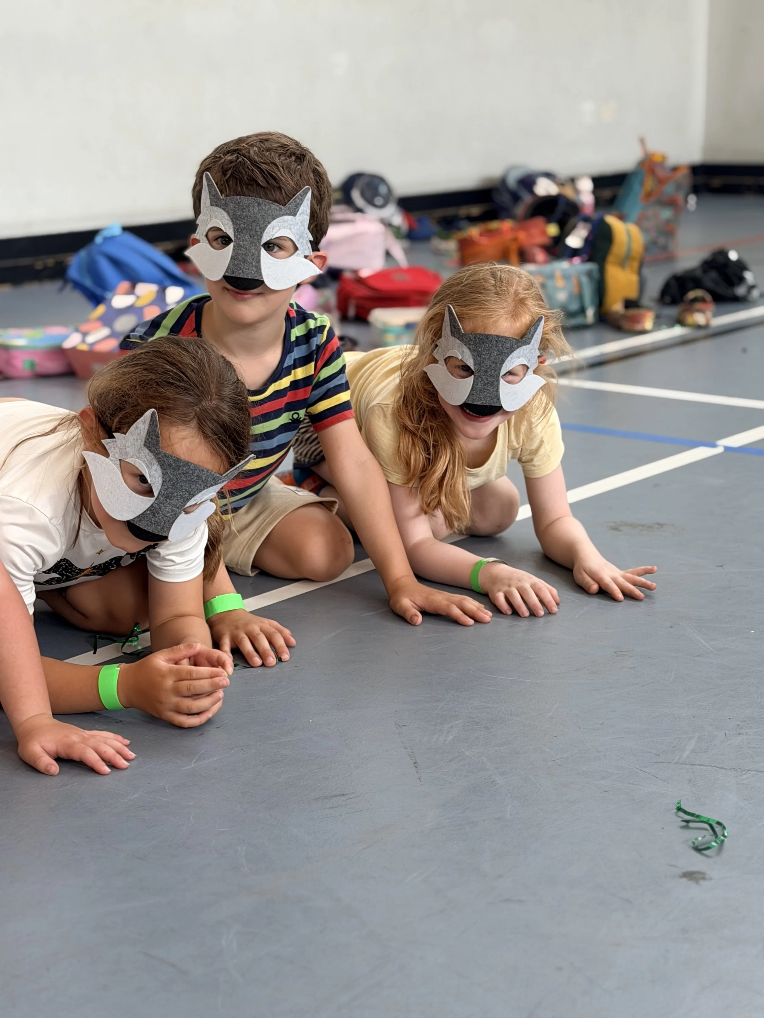 Three children in felt wolf masks crouch on all fours on the sports hall floor, two of them smiling.