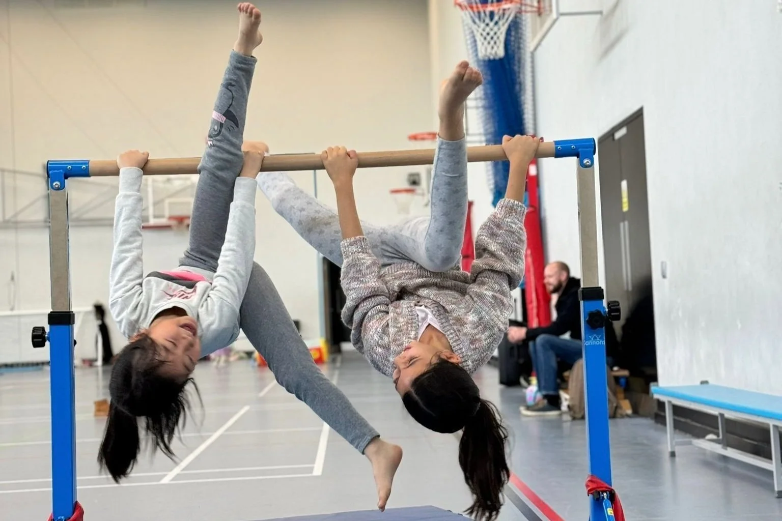 Two smiling girls hang upside down from a low bar in the sports hall.