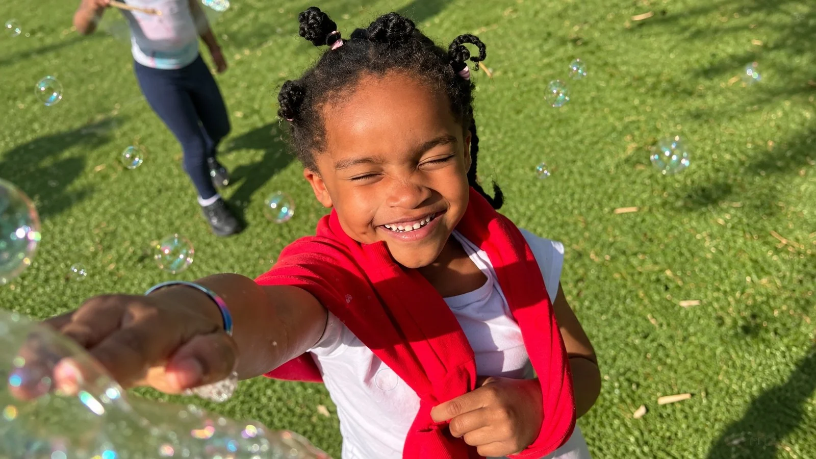 A child laughing and reaching for bubbles outdoors; an edited GymLab Camps photograph.