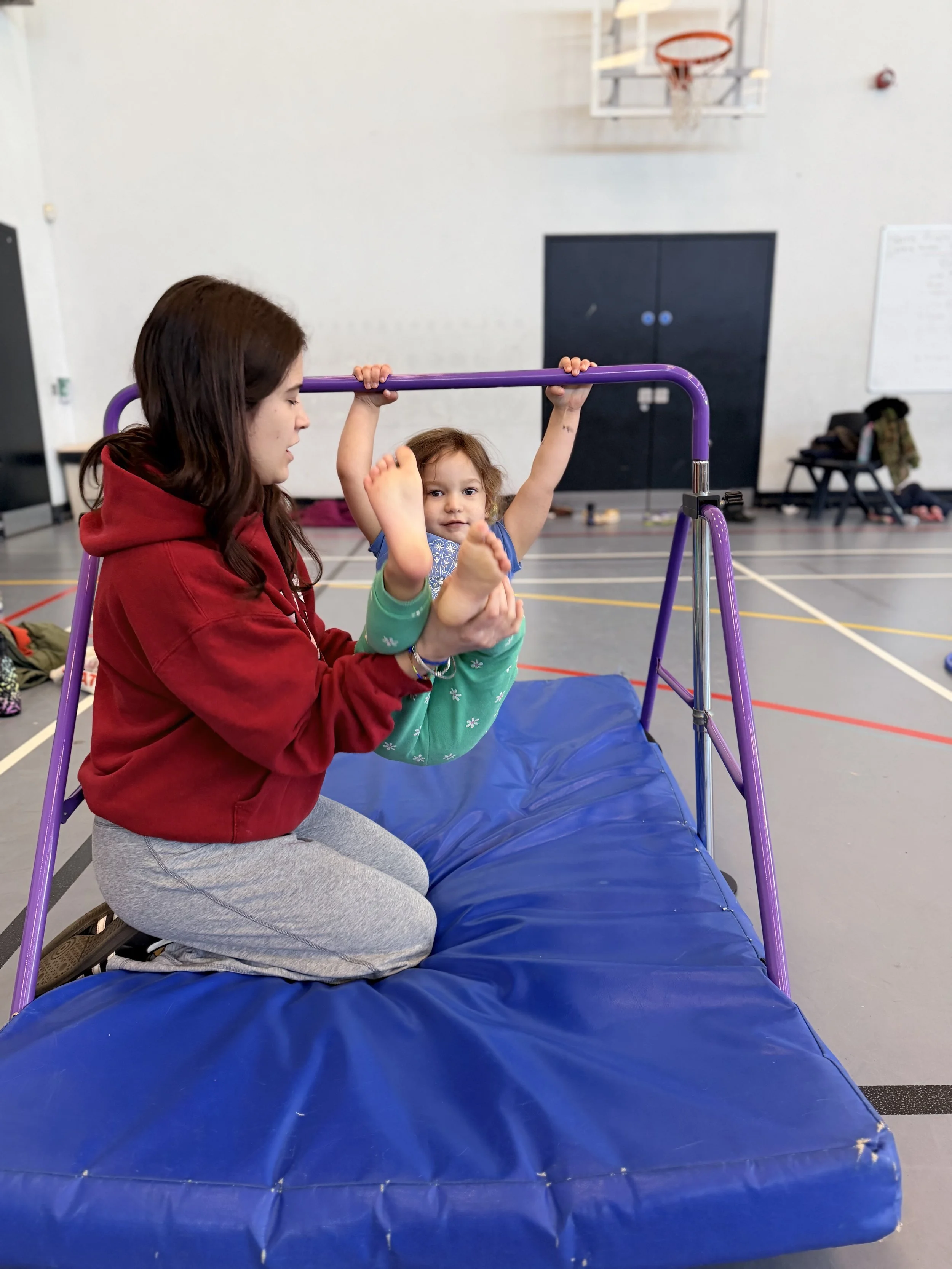 A coach kneels on a soft mat and supports a toddler hanging from a low purple bar.