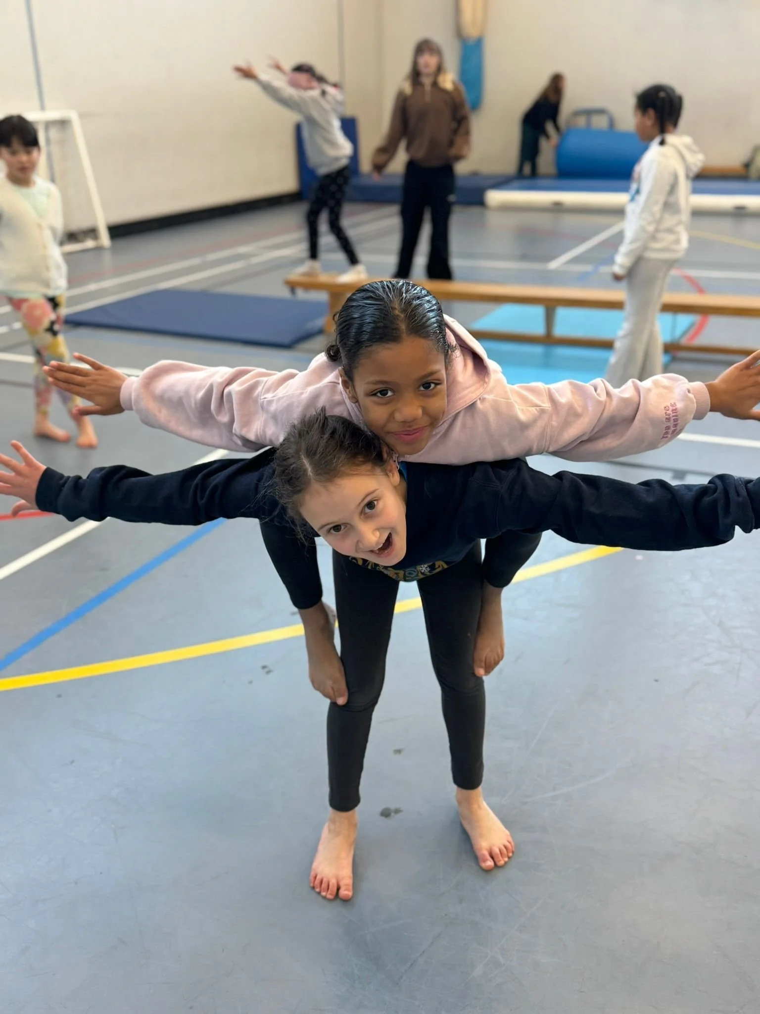 Two girls balance together with their arms spread wide like aeroplanes in the sports hall.